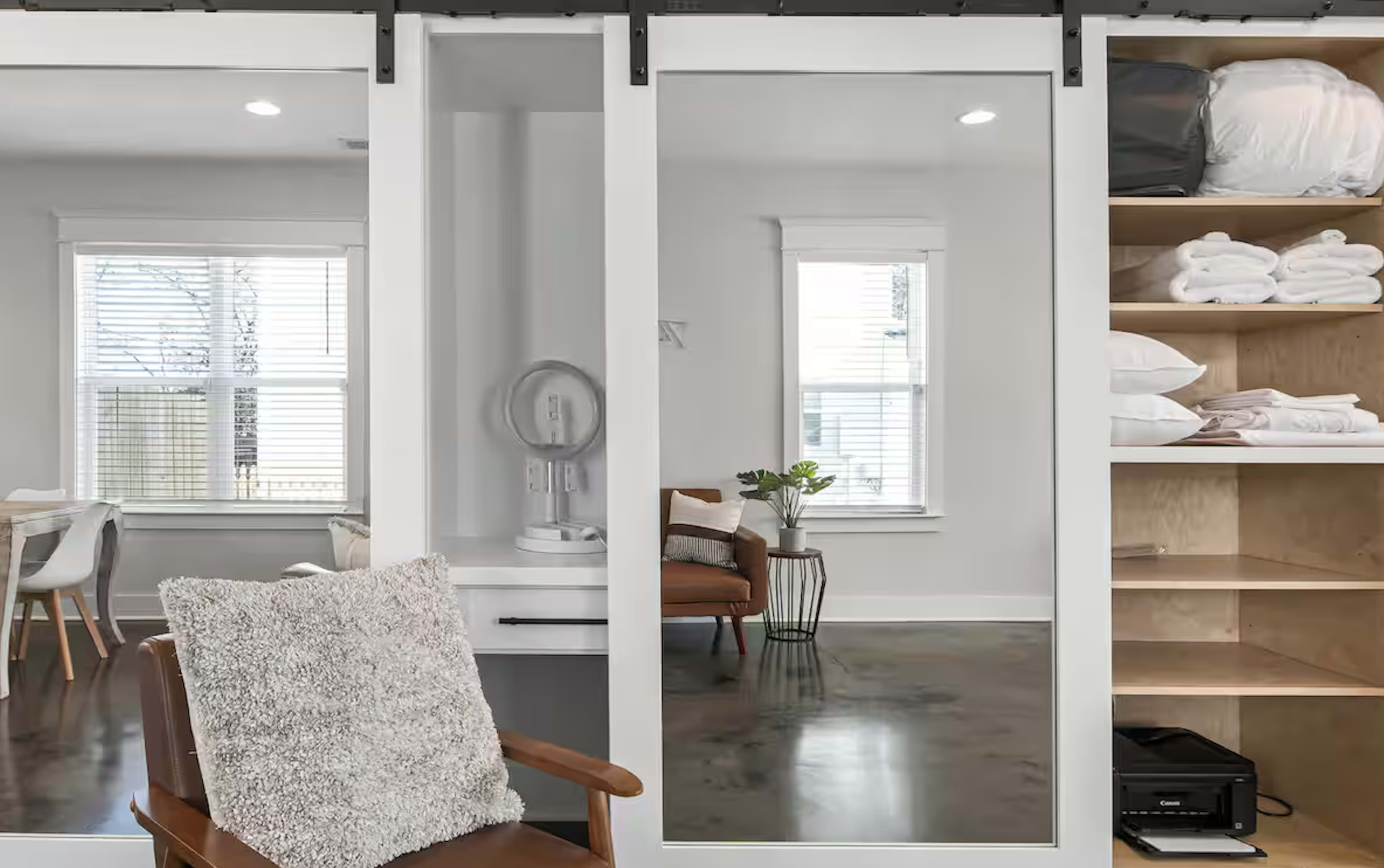 The image shows a modern interior featuring a wooden chair in the foreground, sliding mirrors, a small table with a lamp, and an open shelf with neatly folded towels and a box.