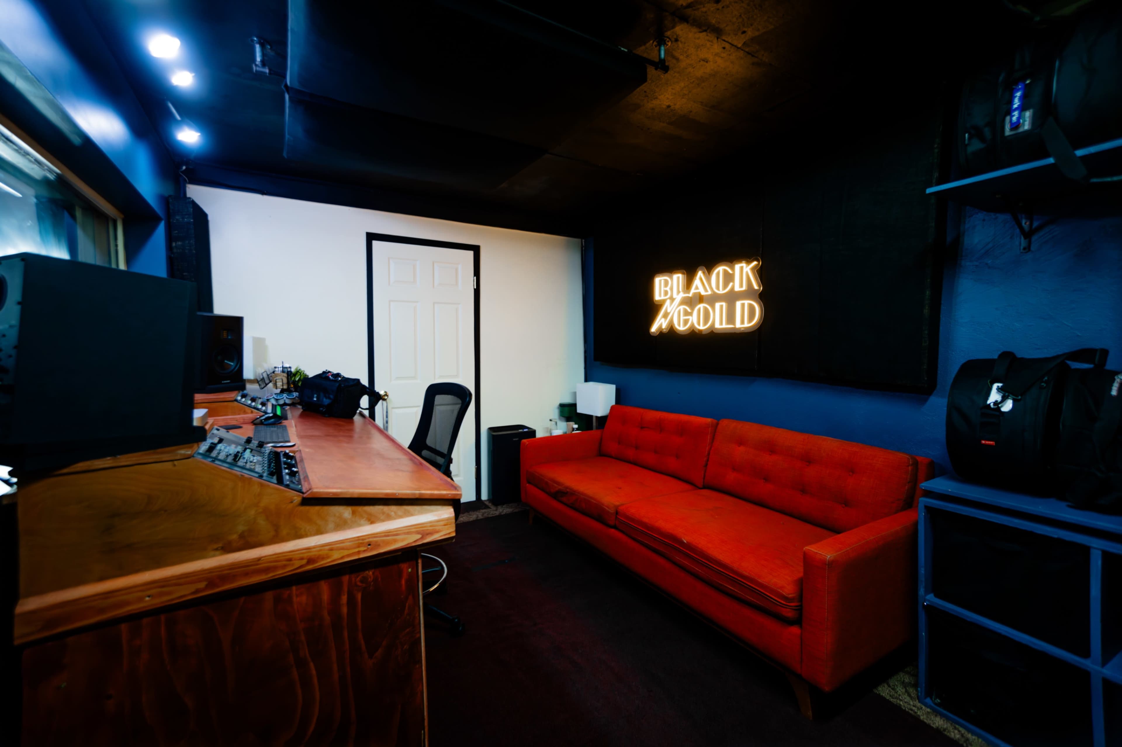 A music studio featuring a red sofa, a wooden desk with audio equipment, and a neon sign that reads "BLACK GOLD."