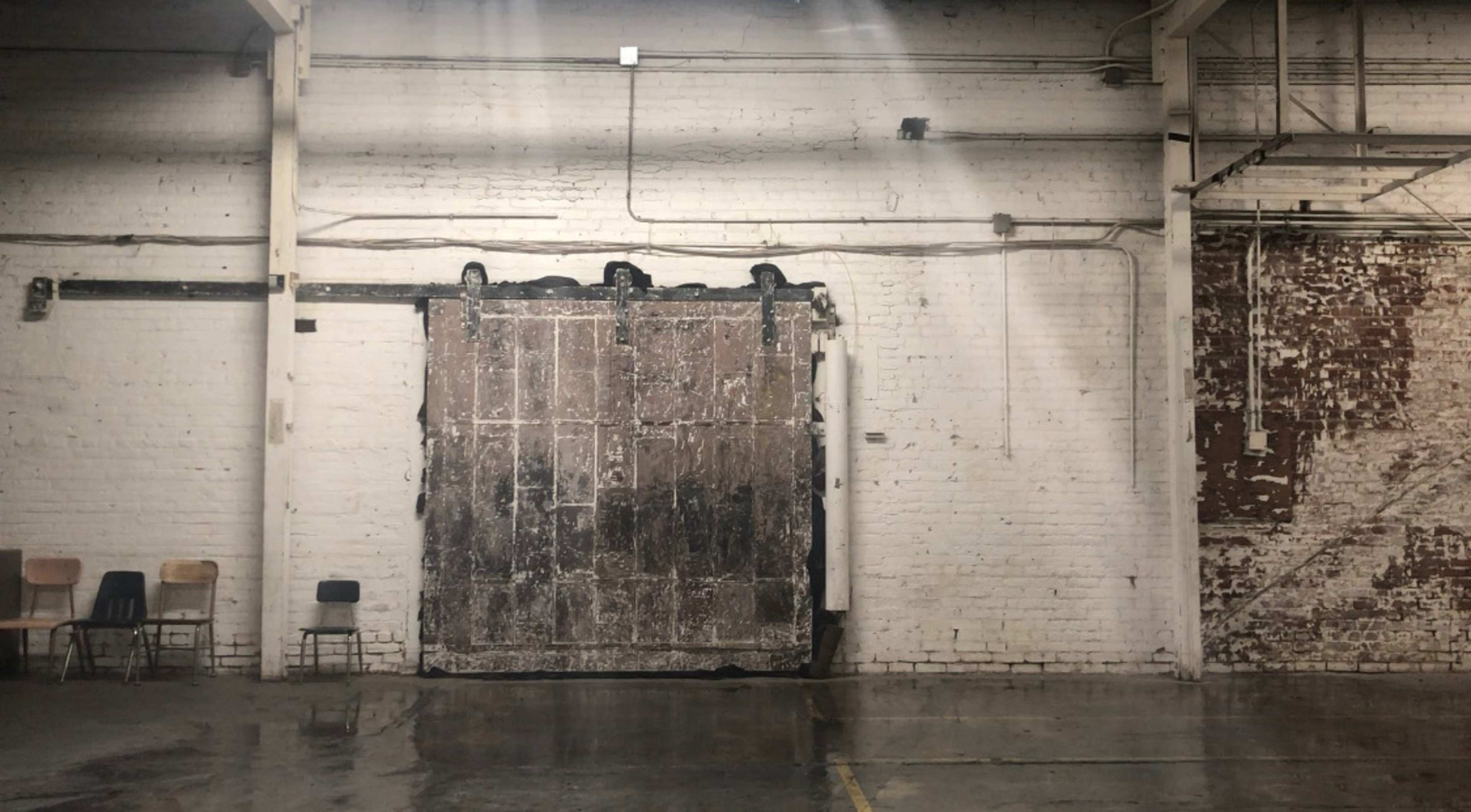 A large, weathered sliding door in an industrial space with exposed brick walls and a polished concrete floor, accompanied by a few empty chairs in the foreground.