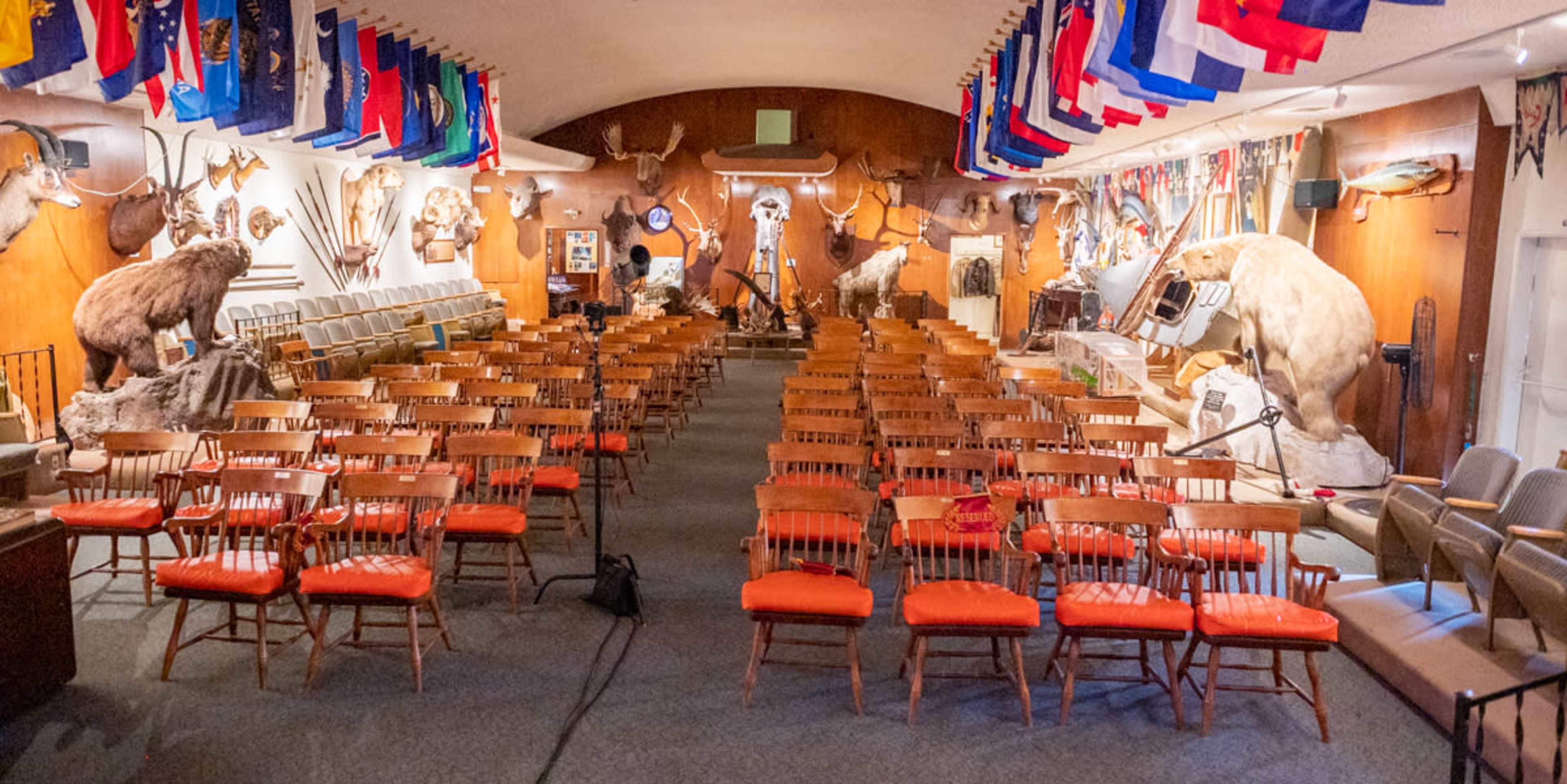 A large room filled with rows of wooden chairs, decorated with mounted animal trophies and flags hanging from the ceiling.