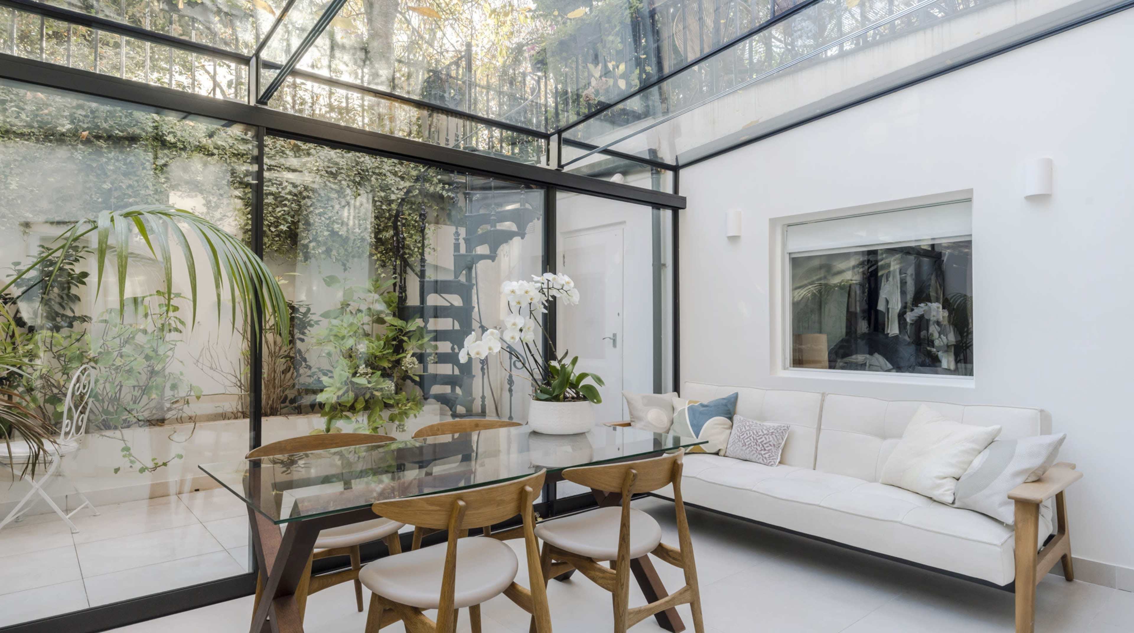 A modern glass-enclosed room featuring a dining table with wooden chairs, a sofa, and indoor plants.