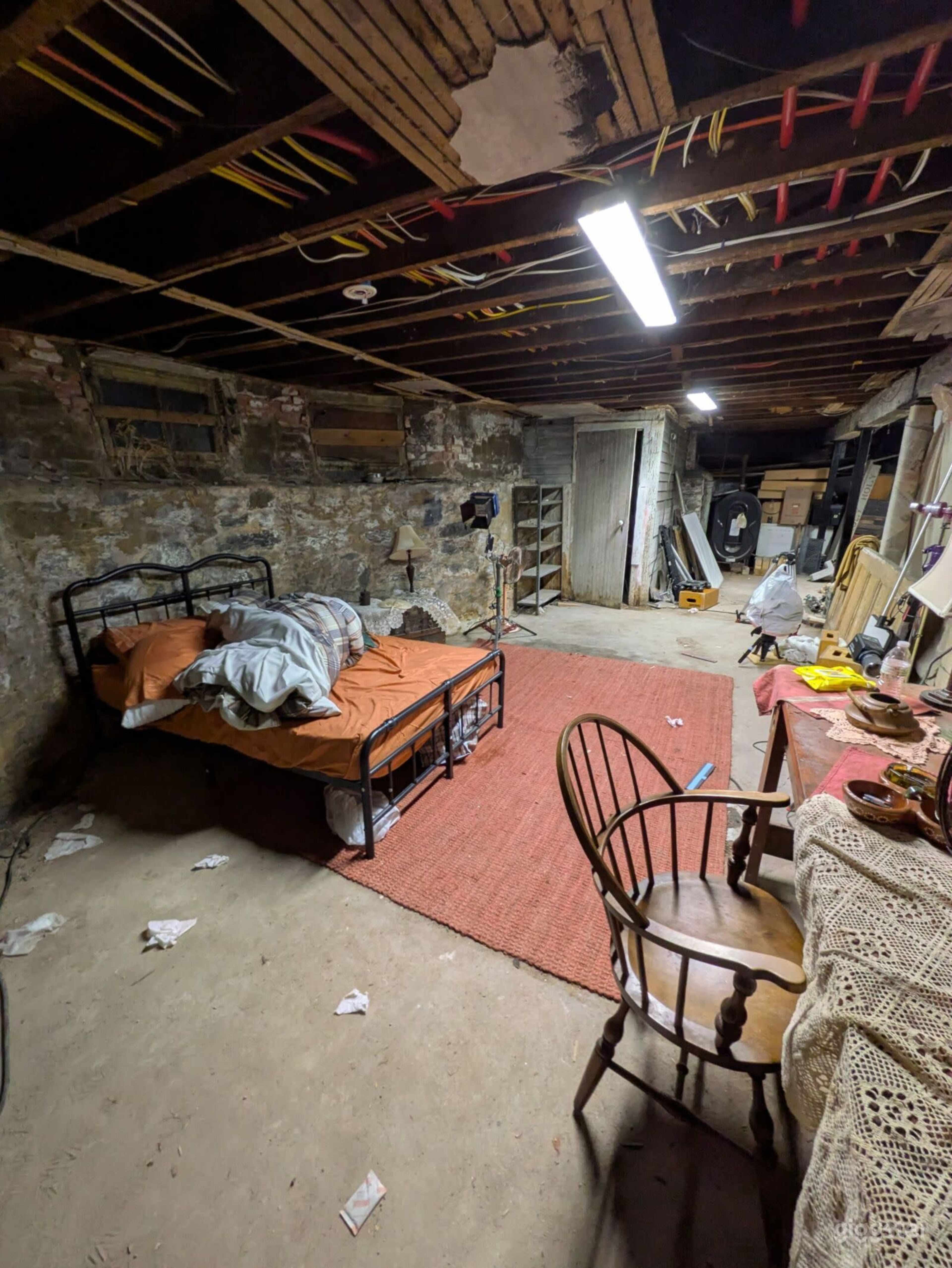 The image shows a cluttered basement with a bed, a chair, and scattered items on the floor.