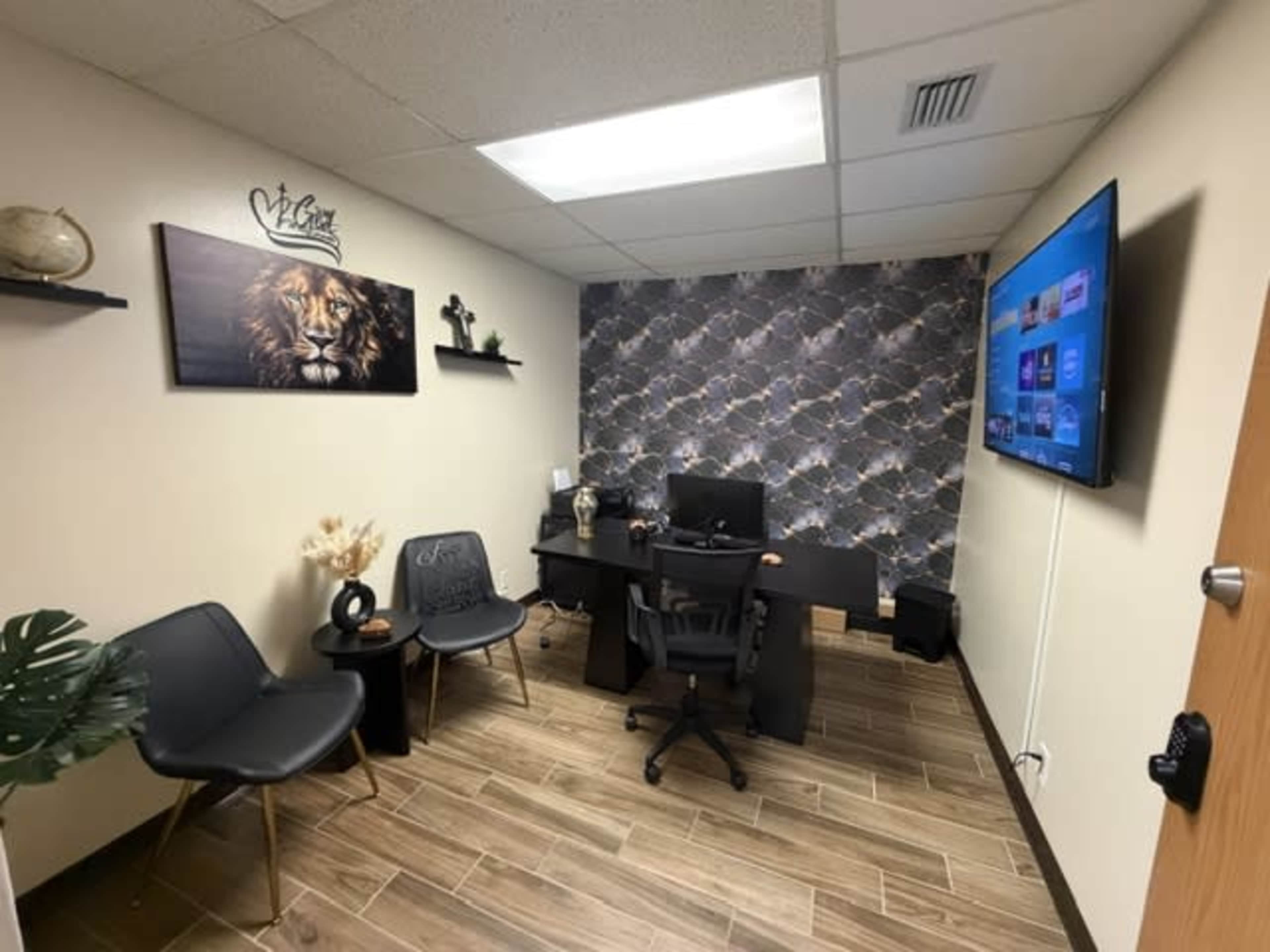 The image shows a small office space with a desk, a computer, two chairs, a decorative wall featuring a lion picture, and a TV mounted on the wall.