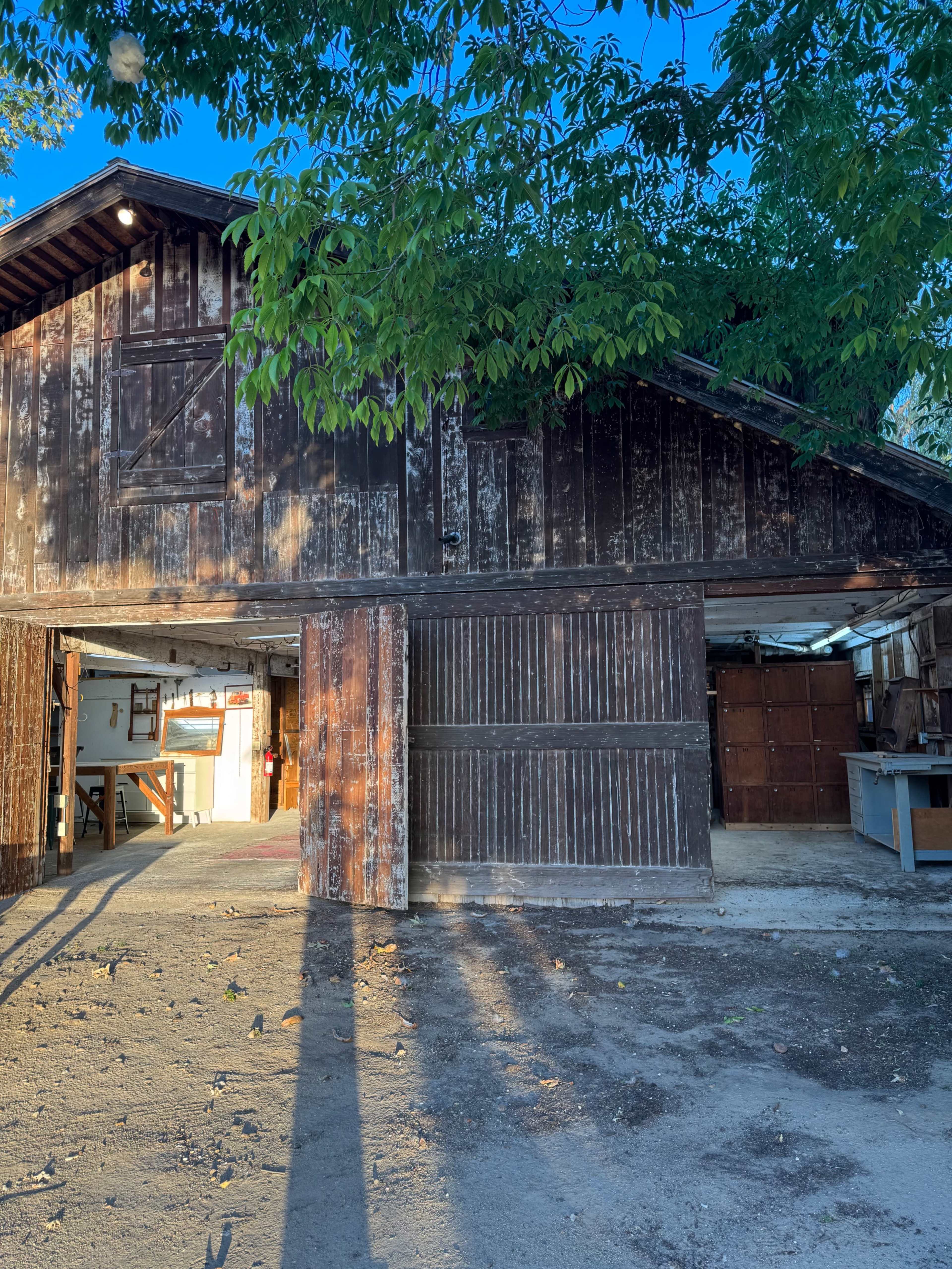 A weathered barn with a large open door stands under a tree in a gravel area.