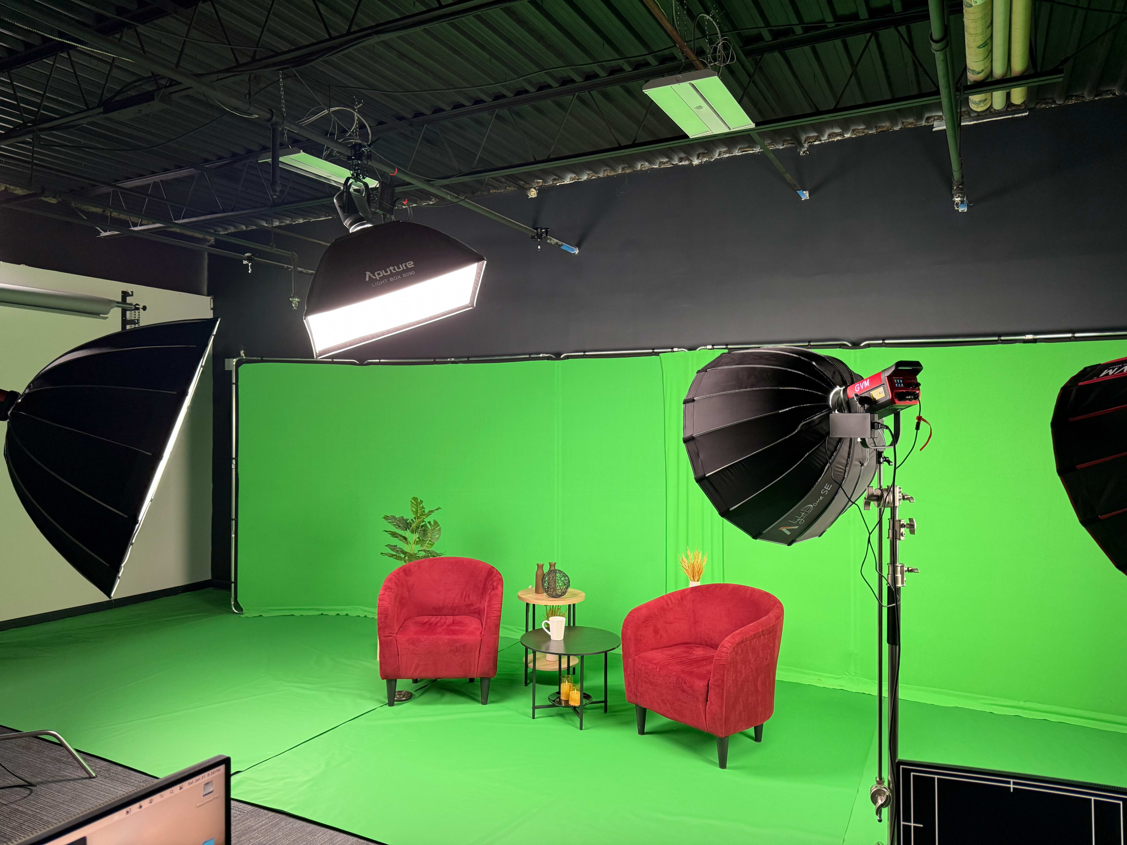 The image shows a studio setup with two red chairs, a small table, and green screen lighting equipment in a room with a green backdrop.