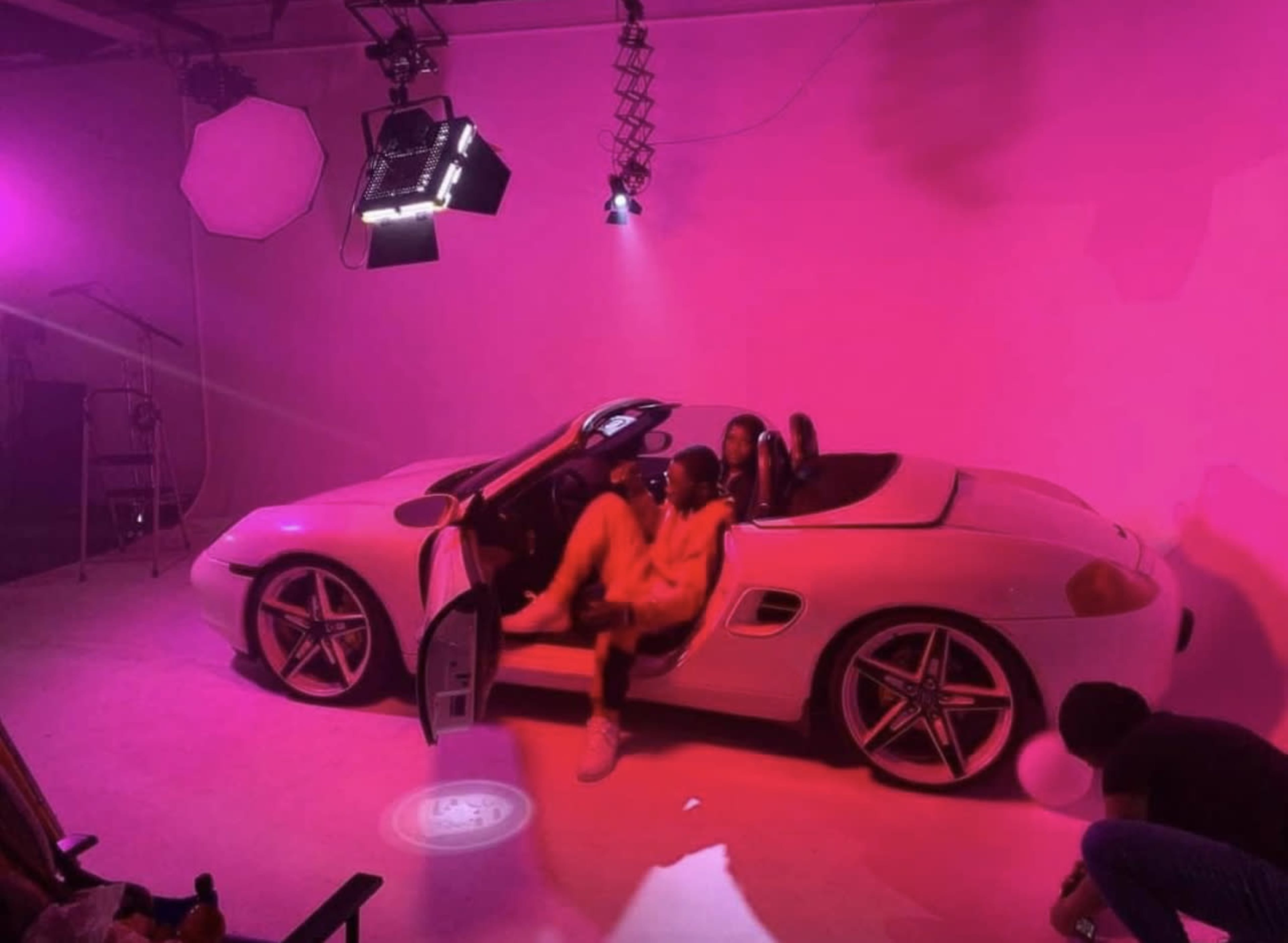 A white convertible sports car is positioned in a studio space illuminated with pink lighting, while a person is exiting the vehicle.
