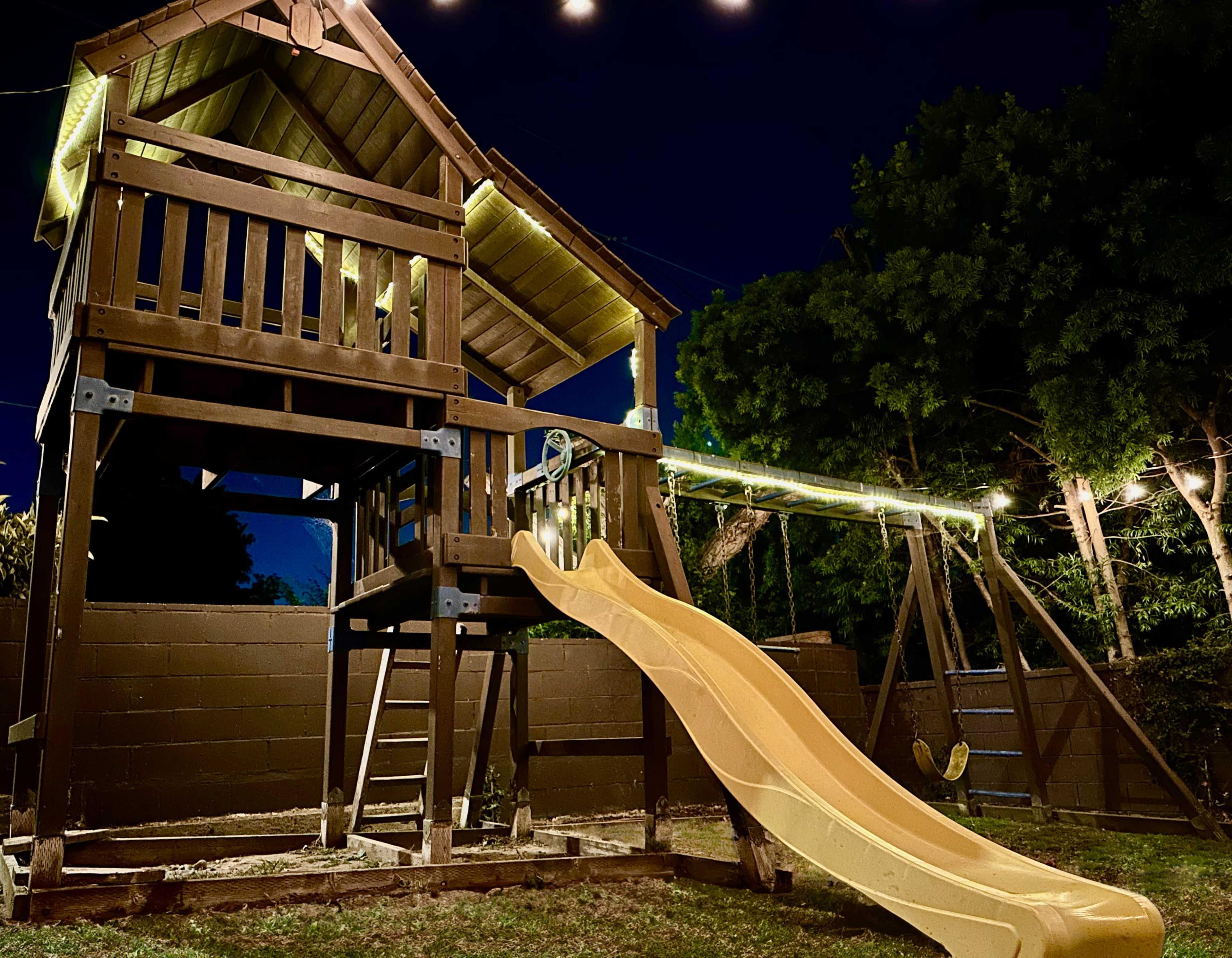 A wooden playset with a slide and swings is illuminated by lights in a backyard at night.