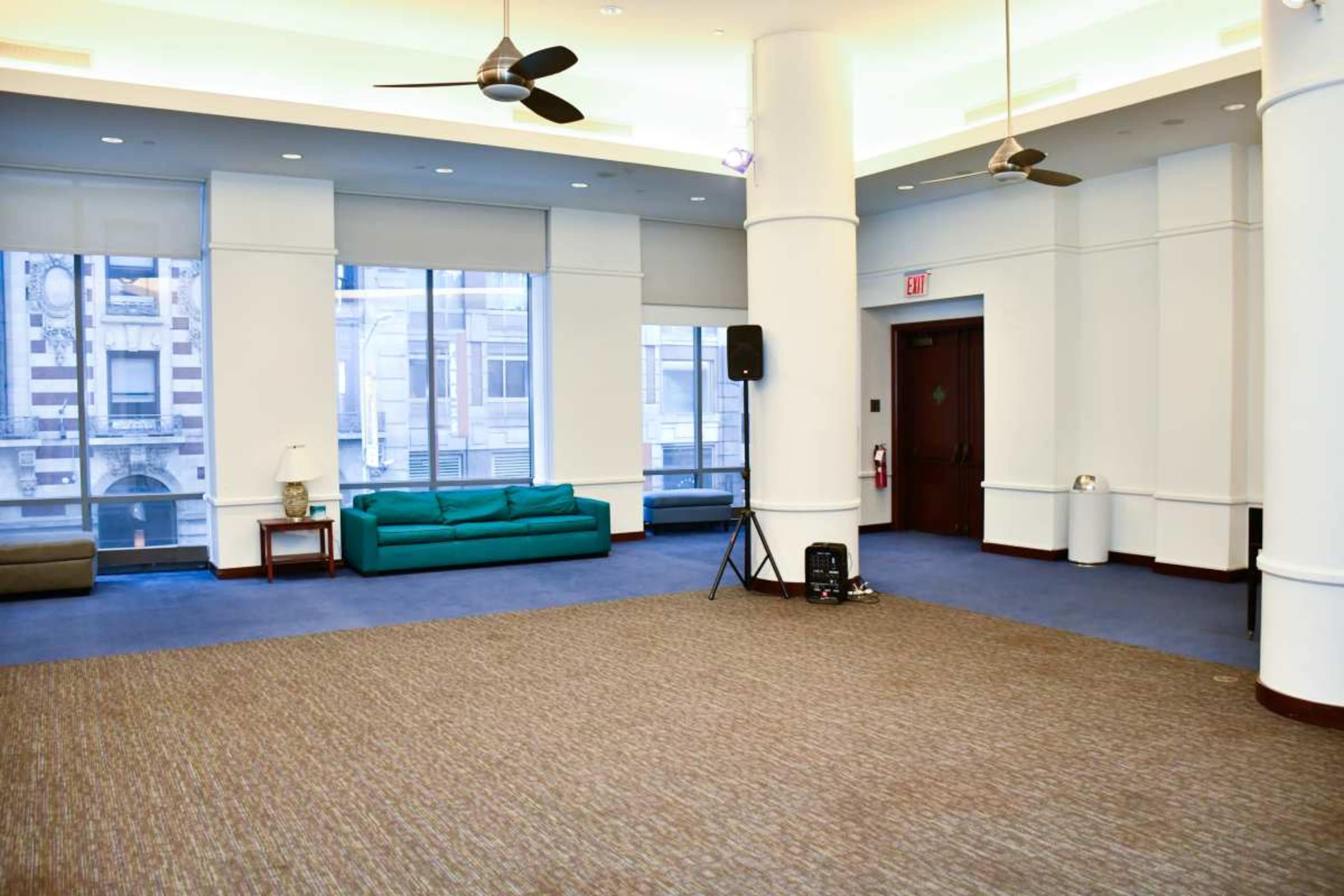 An empty room with light-colored walls, a blue carpet, and large windows, furnished with a green couch and a speaker on a stand.