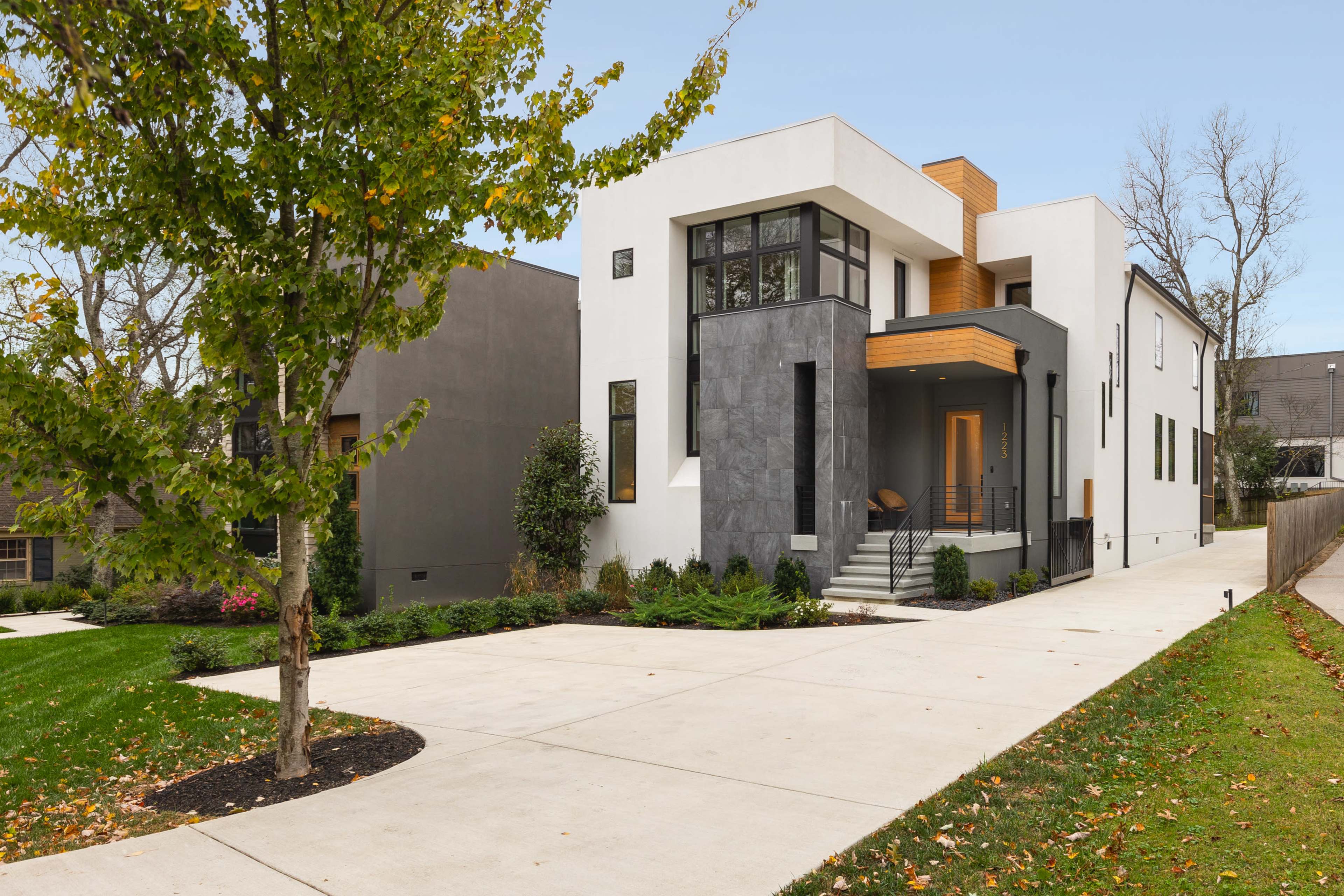 A modern two-story house with a combination of white stucco and dark stone exterior is situated next to a landscaped yard and a paved driveway.