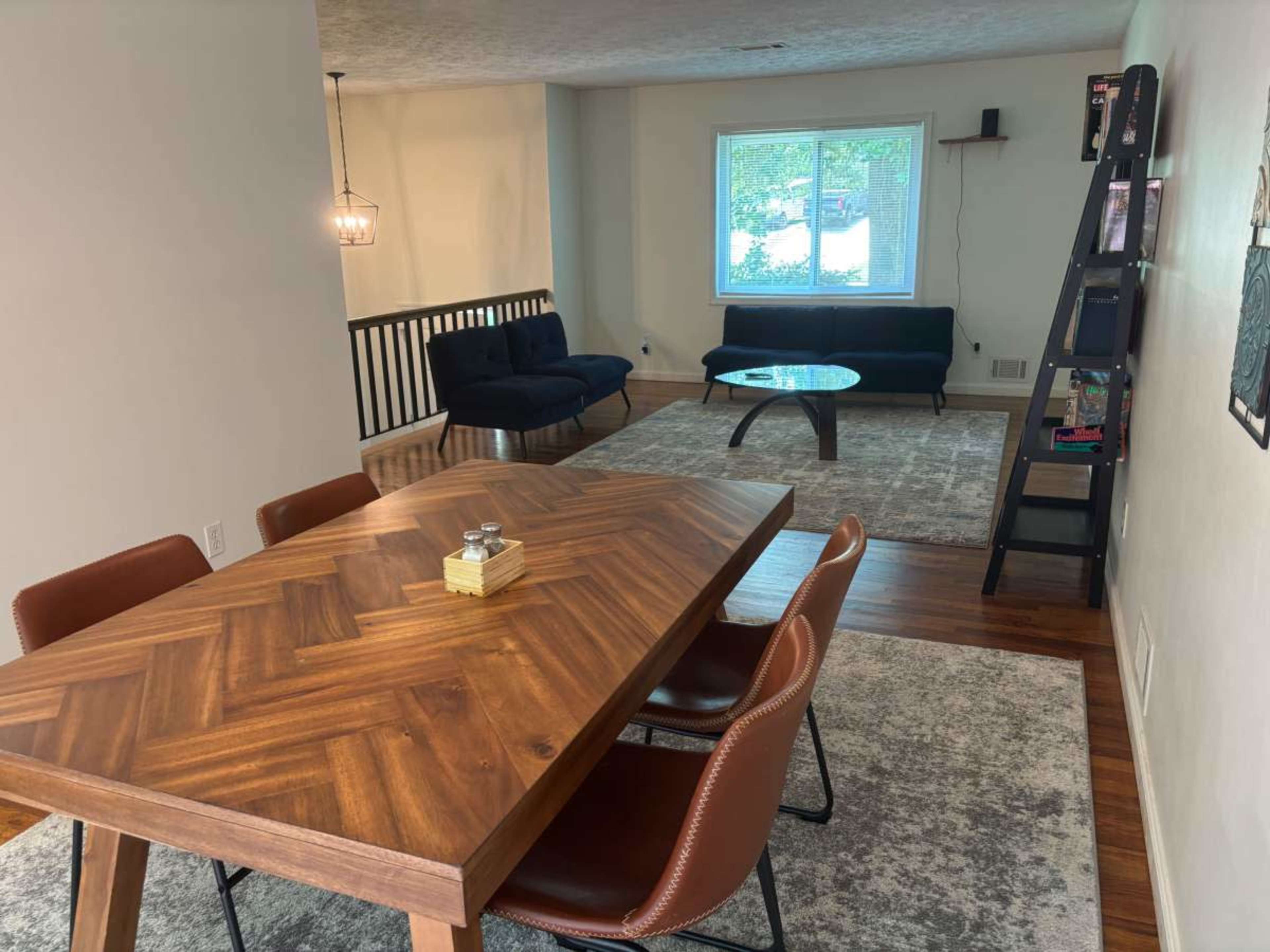 The image shows a dining area with a wooden table and chairs, leading into a living room featuring a sofa and a coffee table, with a window allowing natural light.