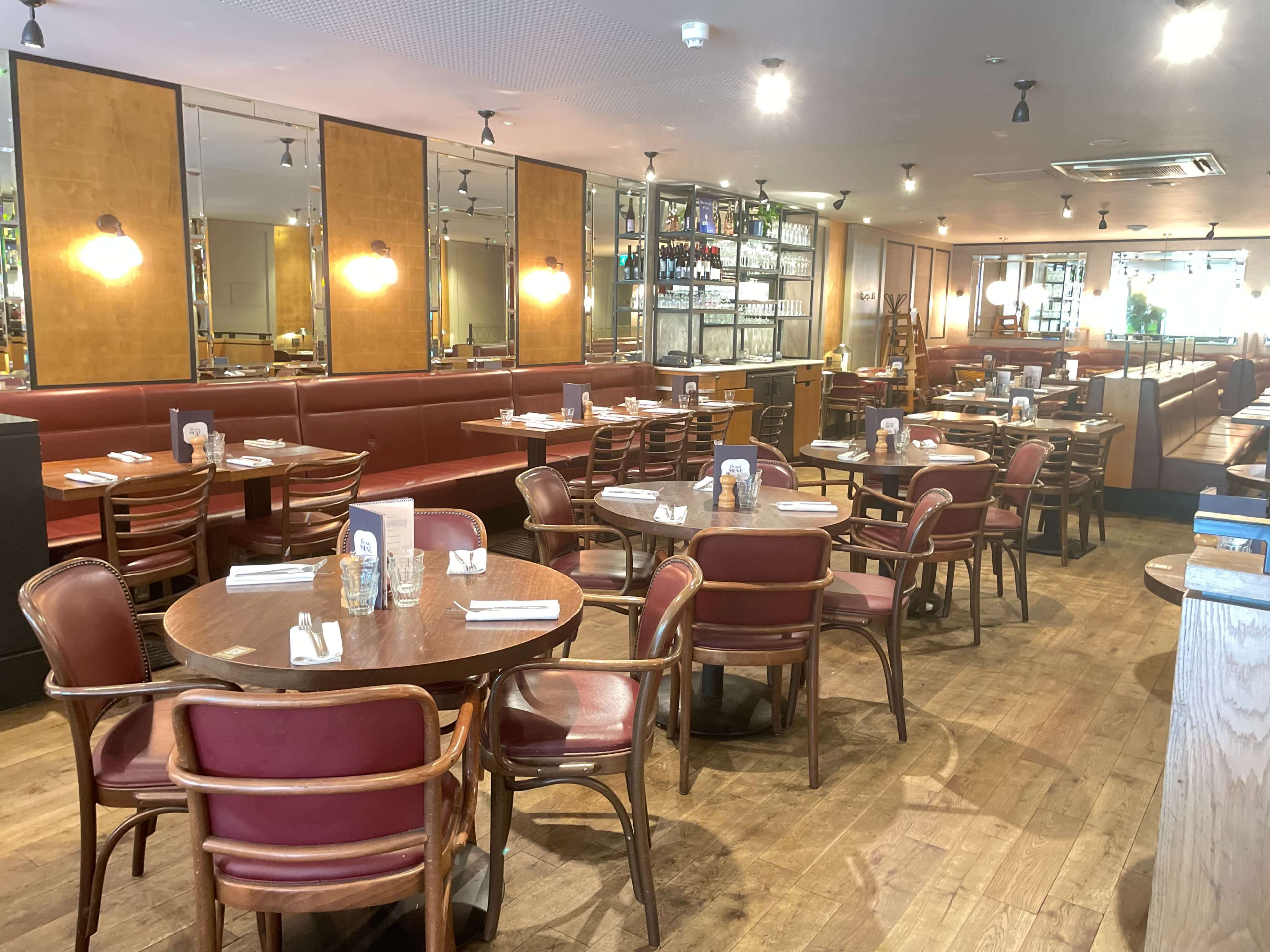 The image shows a spacious restaurant interior featuring wooden floors, red leather booths, and circular wooden tables, all arranged neatly.
