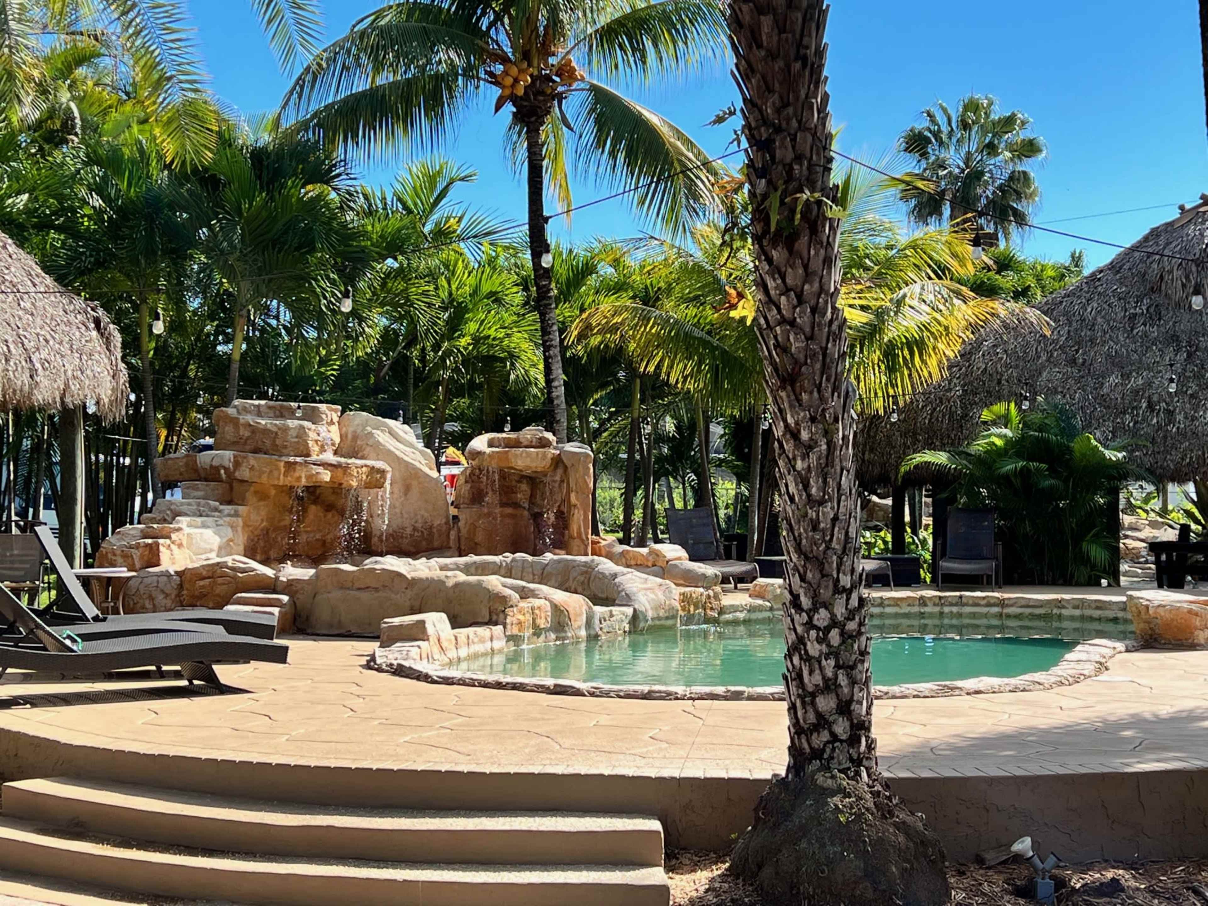 A swimming pool with a rock formation and waterfall is surrounded by palm trees and thatched-roof cabanas.