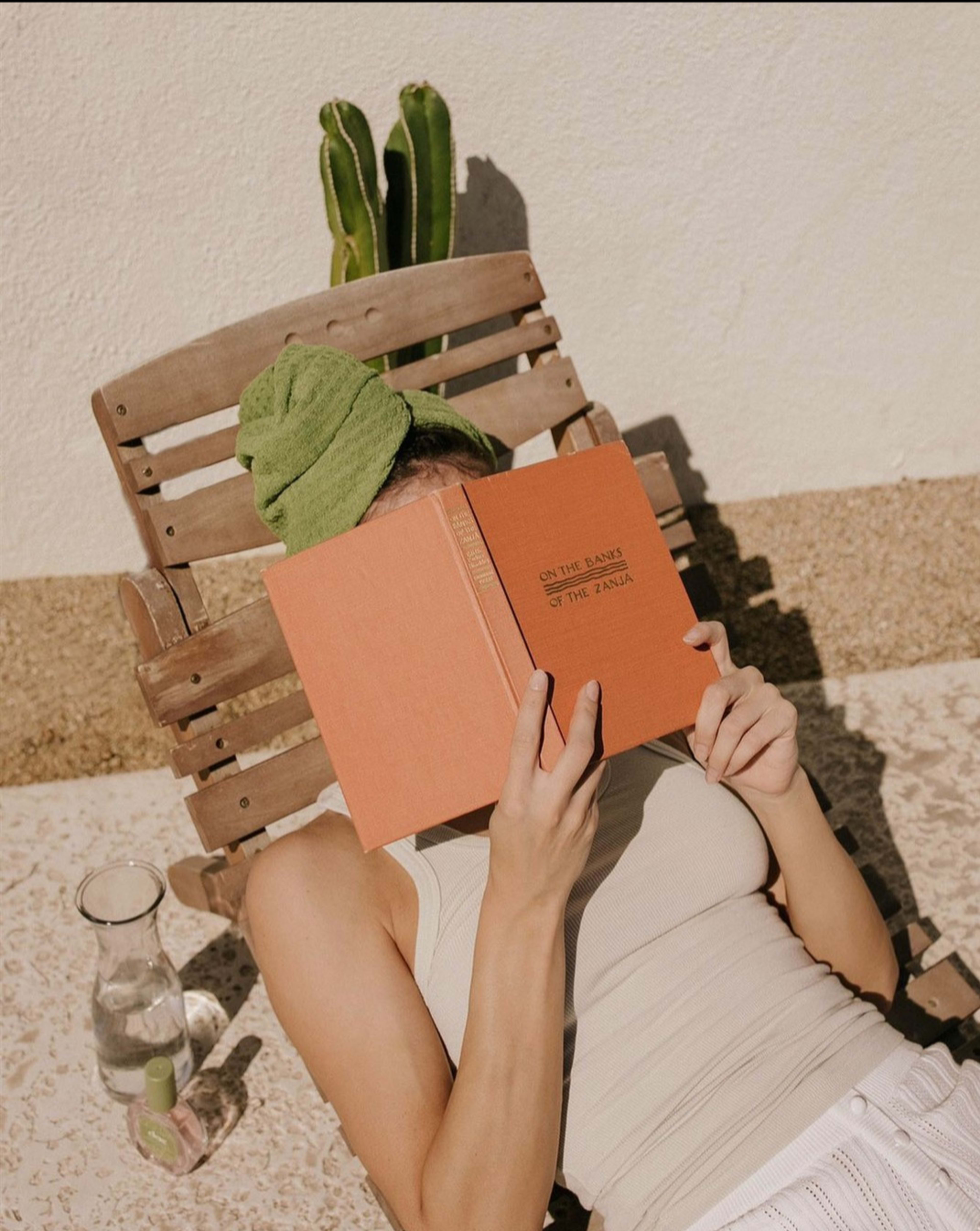 A person lies on a wooden lounge chair, reading a book titled "On the Banks of the Nile," with a green towel on their head and a glass of water beside them.