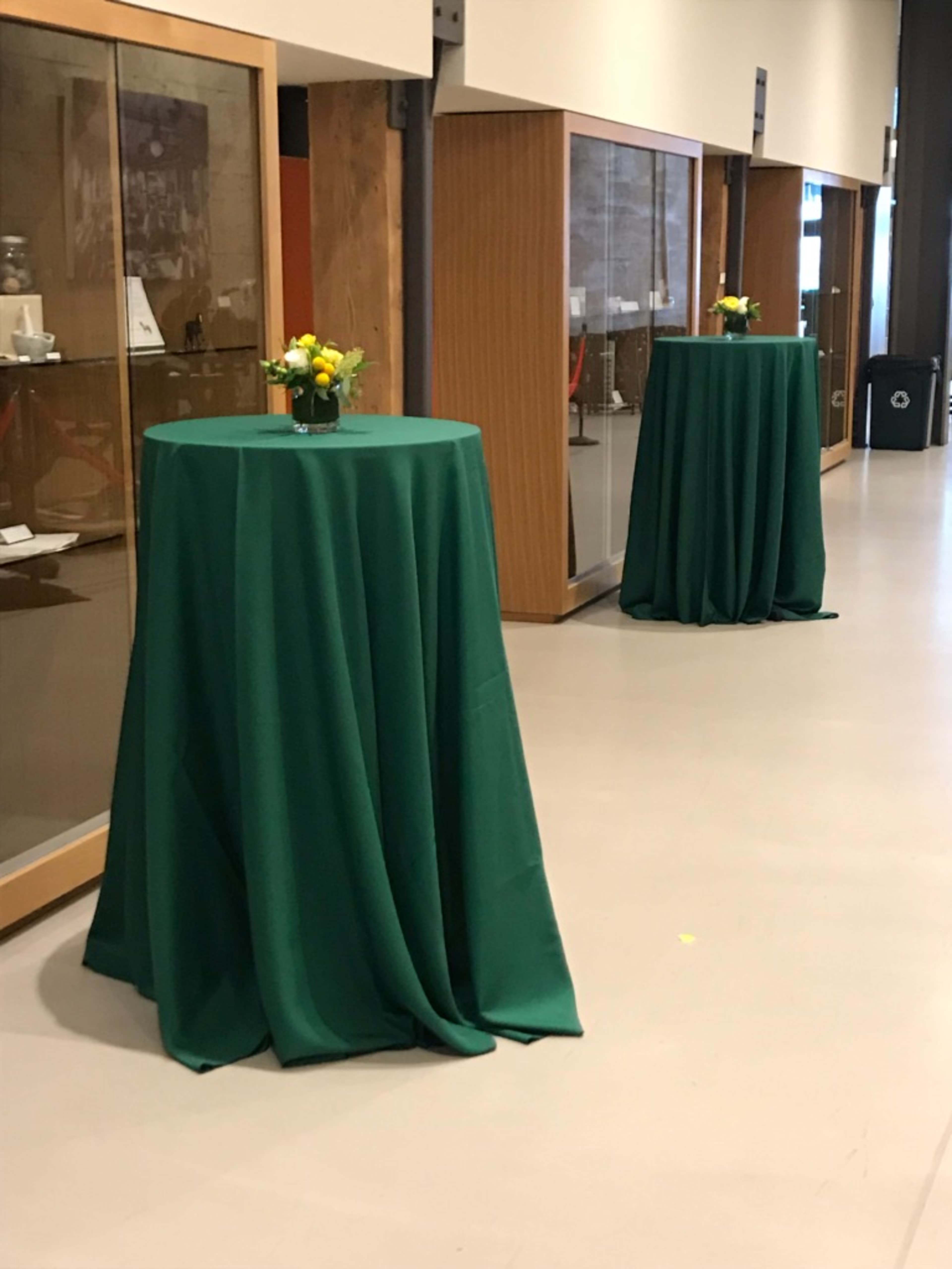 Two tall tables covered with green tablecloths, each adorned with a small flower arrangement, positioned in a hallway with display cabinets in the background.