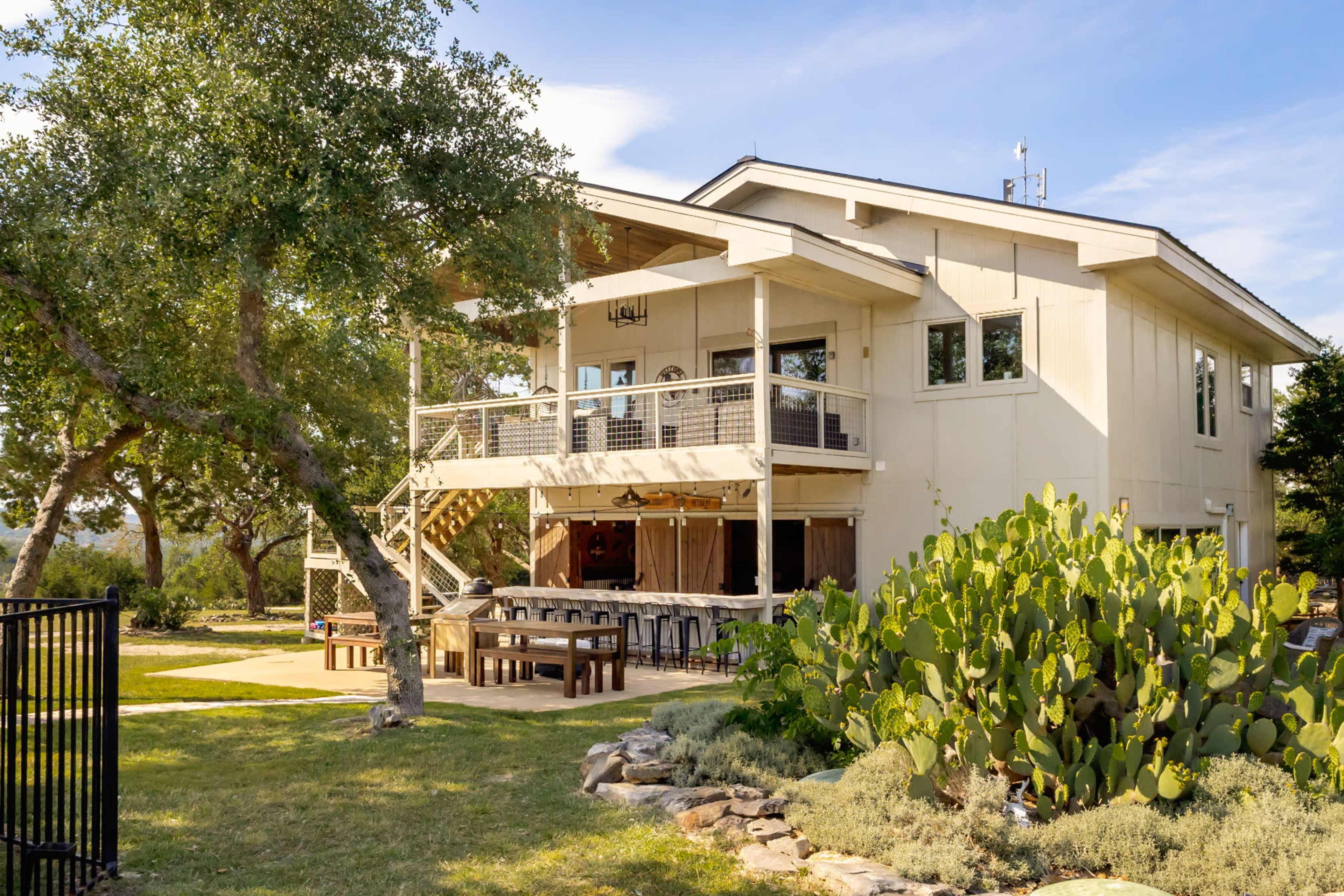 The image shows a modern two-story house with a spacious deck, surrounded by trees and a cactus garden.
