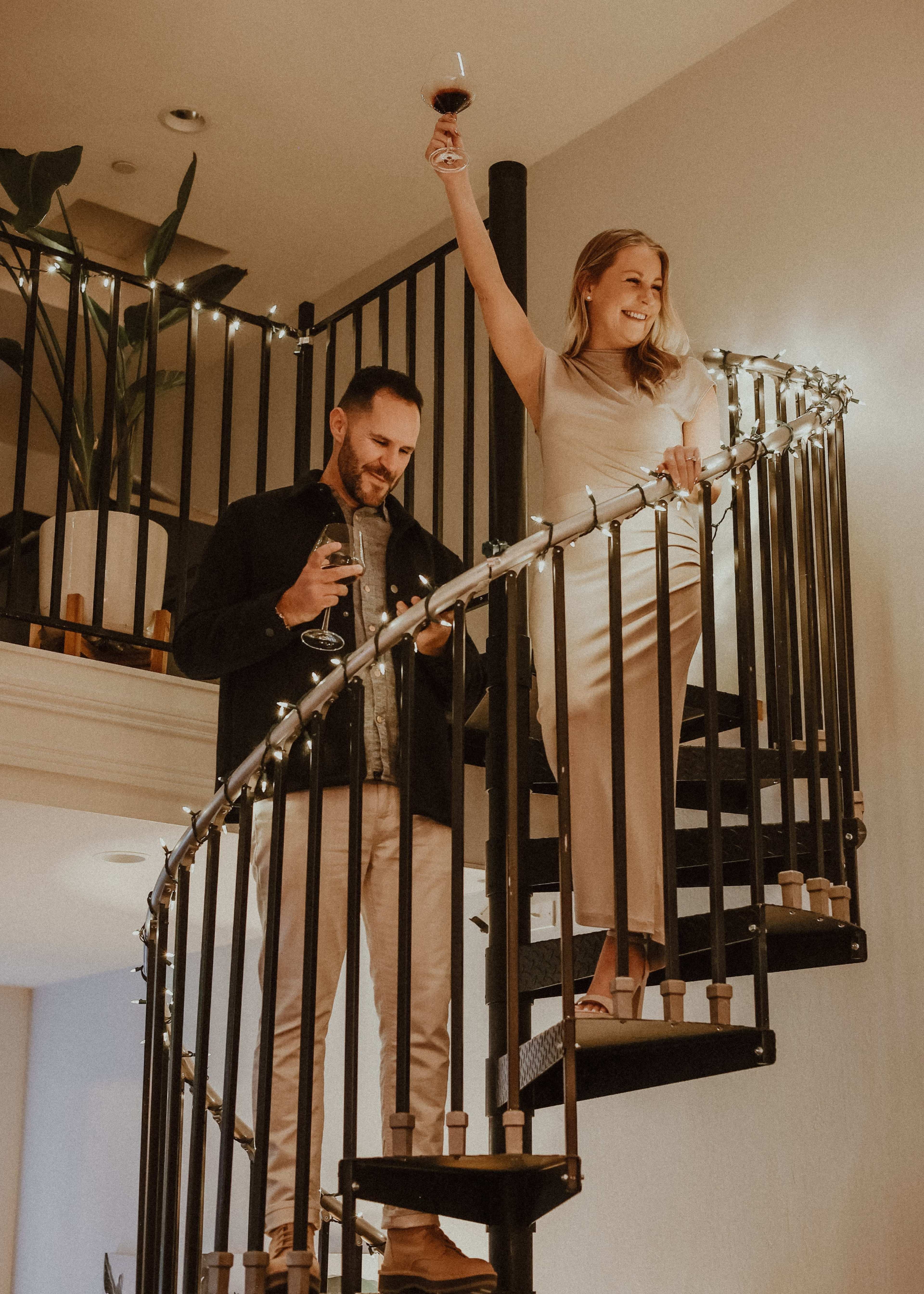 A man stands on the lower step of a spiral staircase holding a glass, while a woman above raises her glass in celebration.