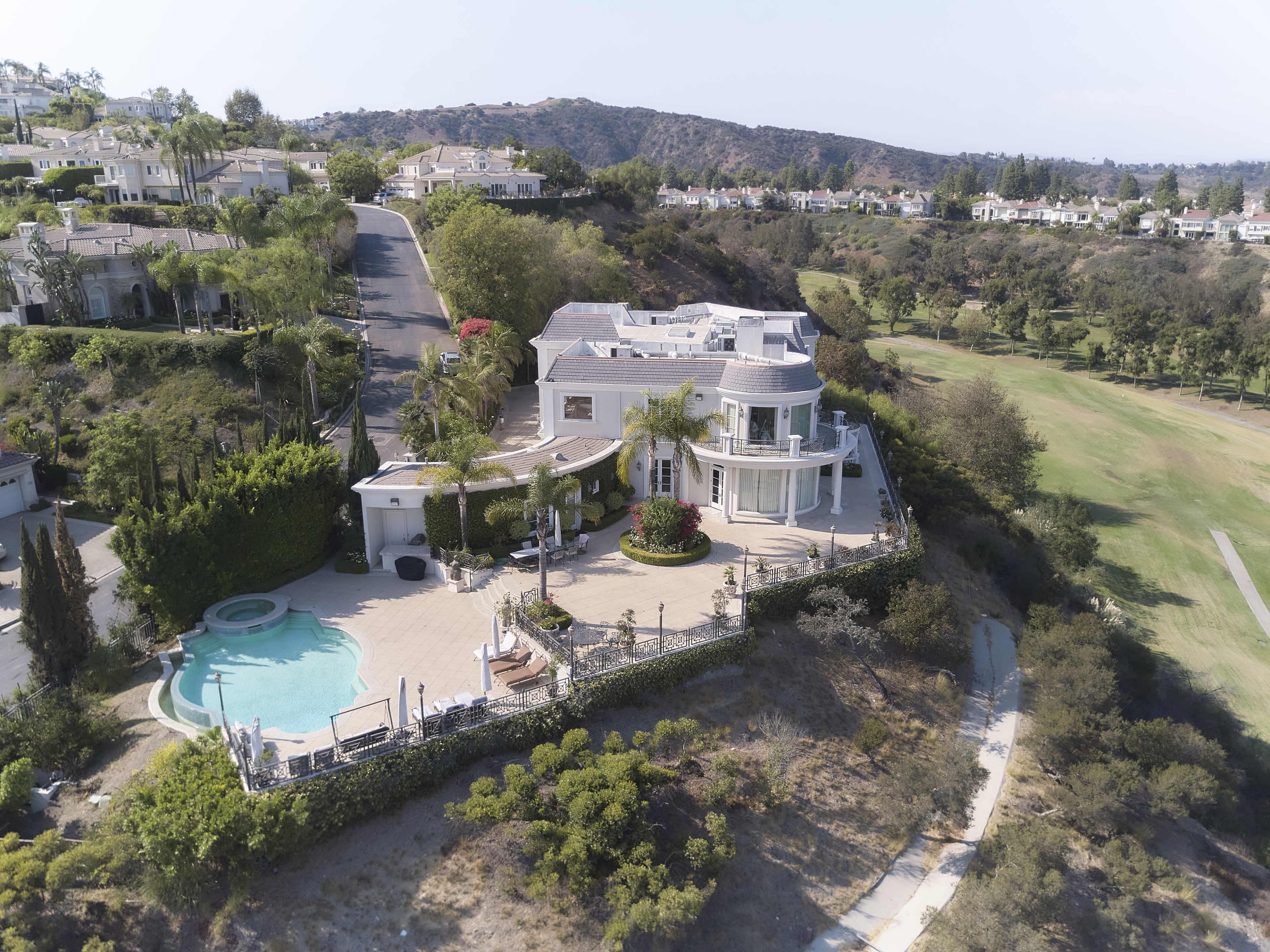 A modern house with a curved design sits on a hillside overlooking a golf course, featuring a pool and landscaped gardens.