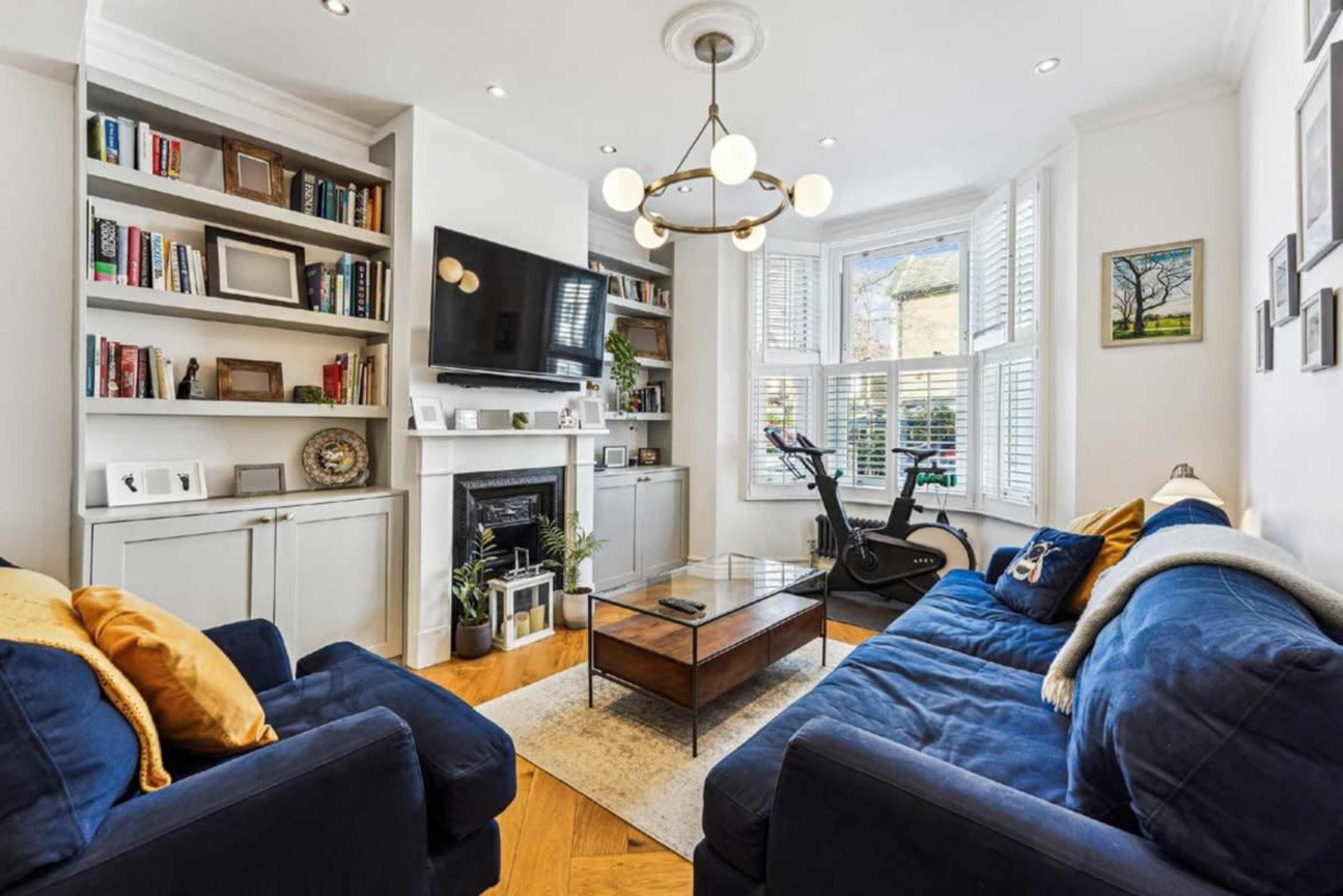 The image shows a modern living room with a blue sofa, a wooden coffee table, a wall-mounted television, and shelves filled with books and decorative items.