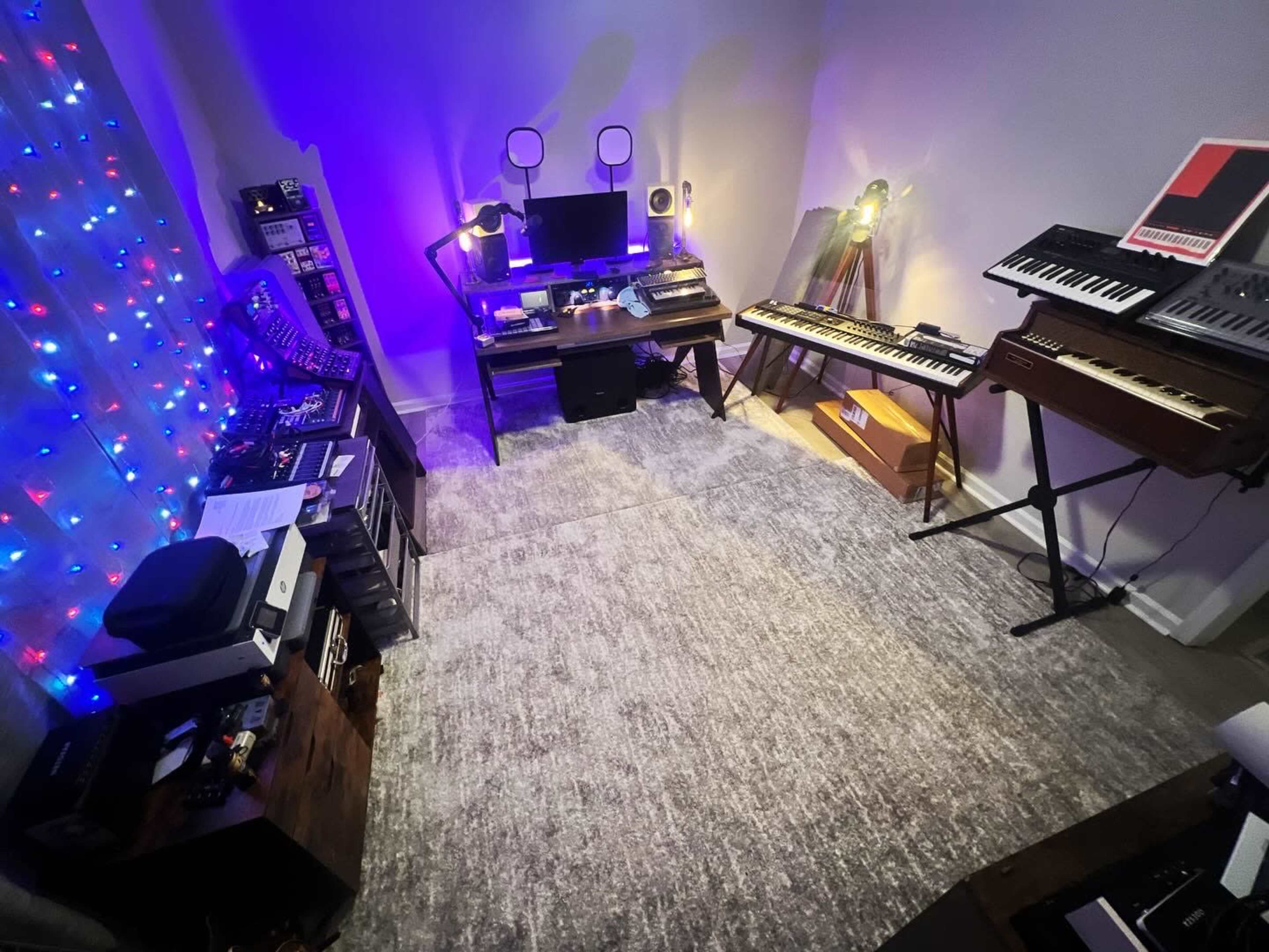 A music studio featuring electronic instruments, a desk with computers and monitors, and colorful lights along the wall.
