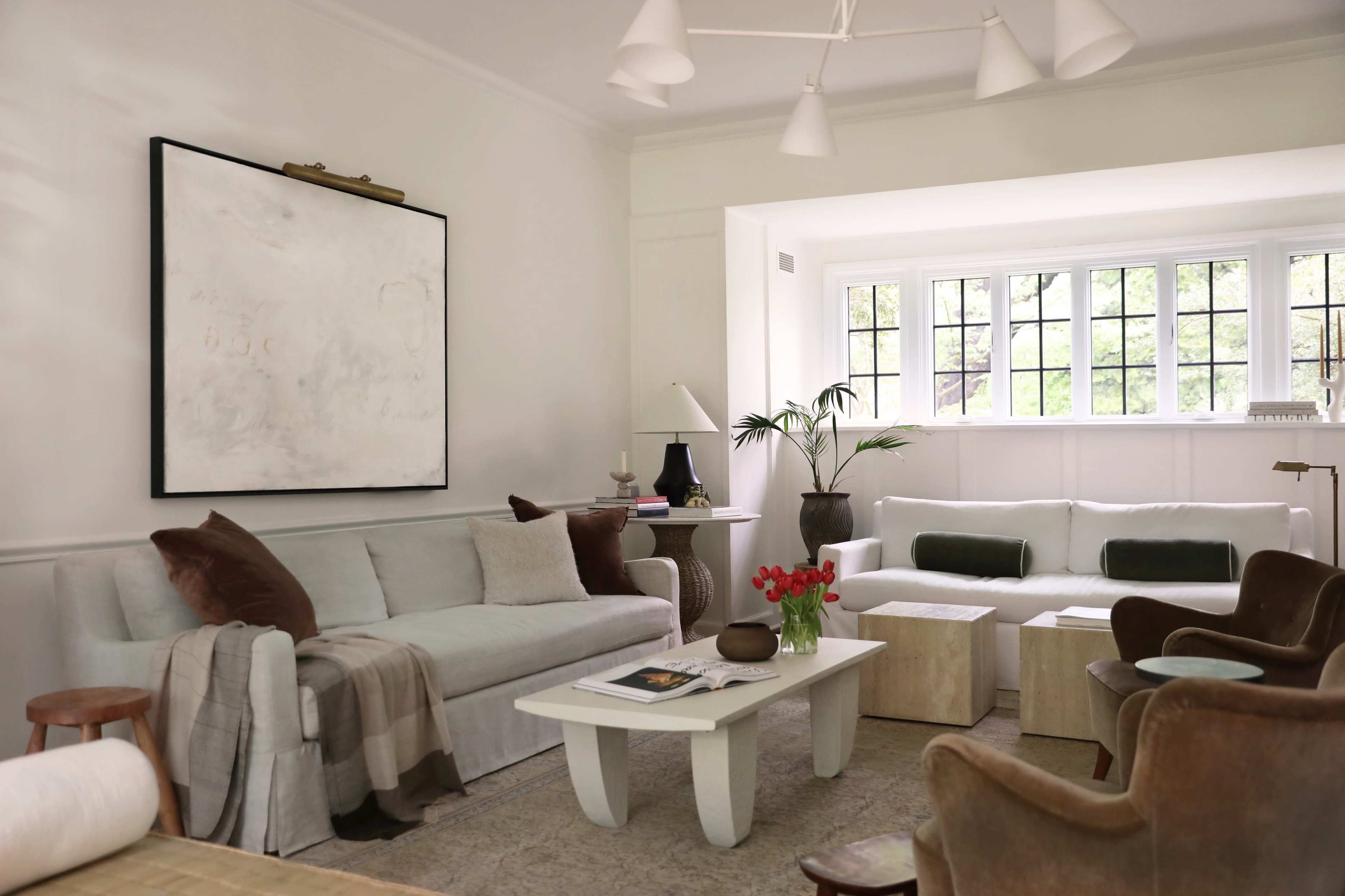 The image shows a modern living room with light-colored furniture, a large abstract painting on the wall, and large windows allowing natural light.