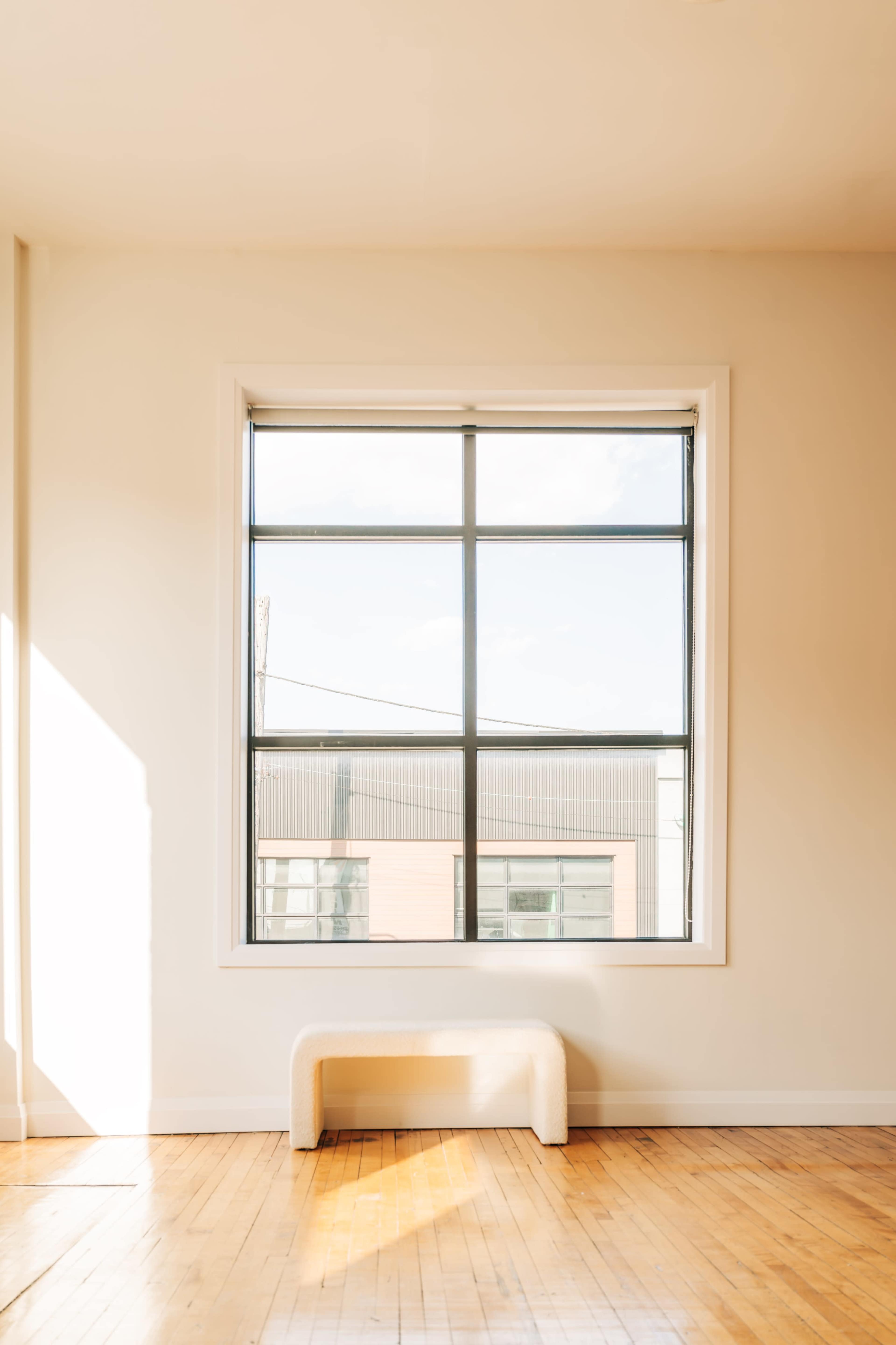 A large window with a grid design allows sunlight to enter a minimalist room featuring a simple bench in front of it.