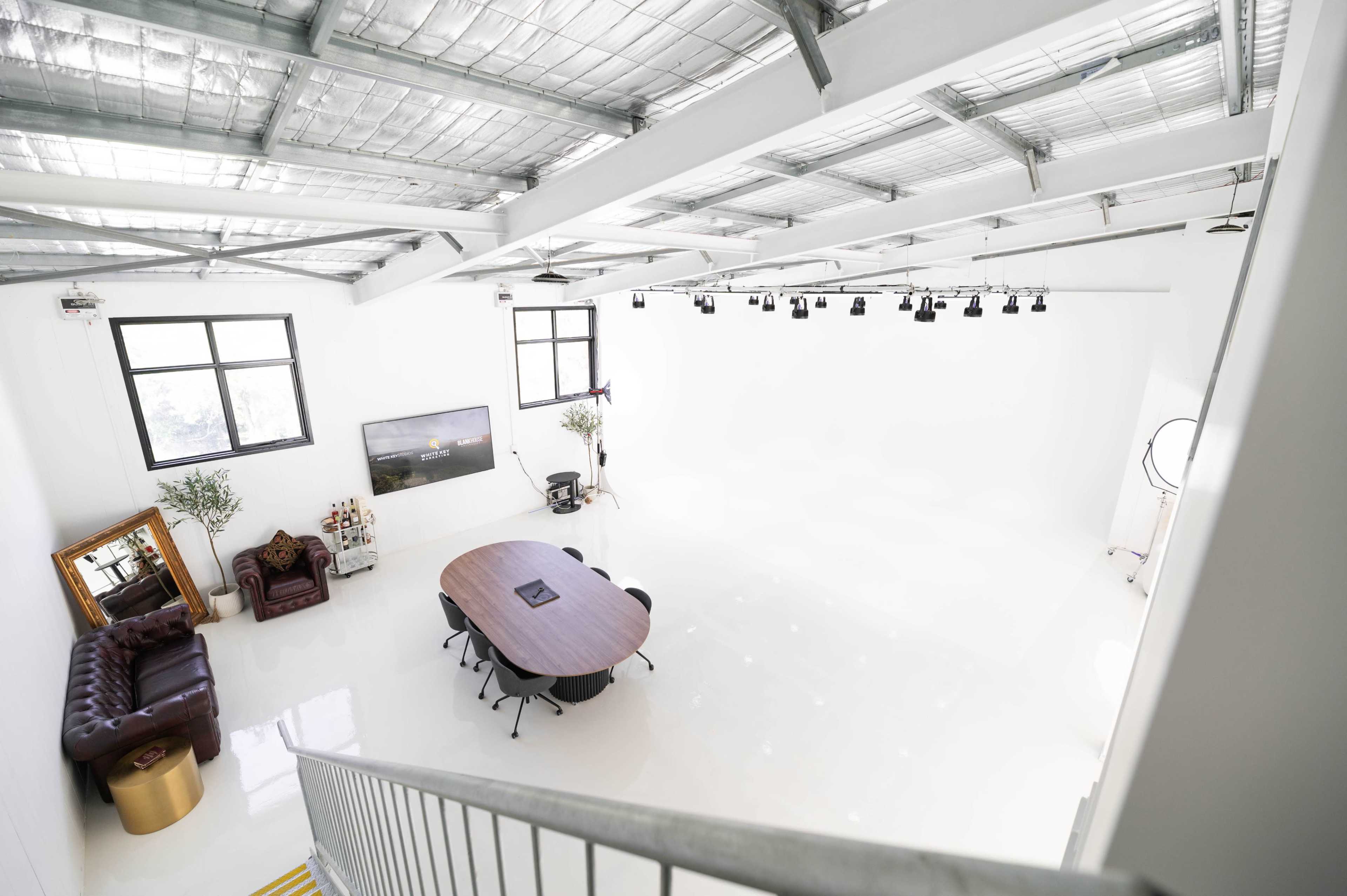 The image shows a spacious, brightly lit studio with a large round table surrounded by black chairs, a leather couch, and a flat-screen TV against one wall.