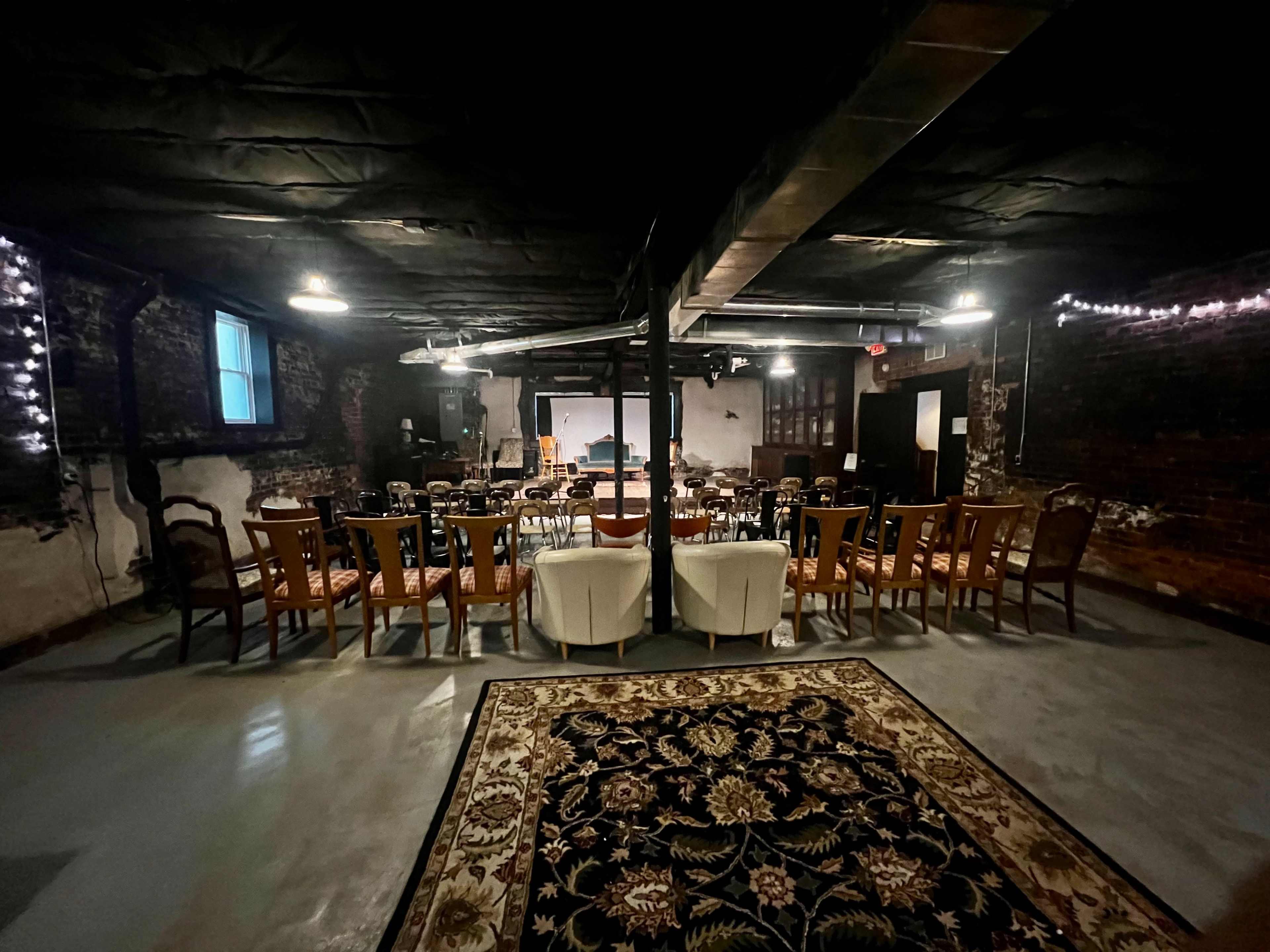 The image shows a dimly lit basement room with rows of chairs arranged around a central seating area, decorated with a large rug.