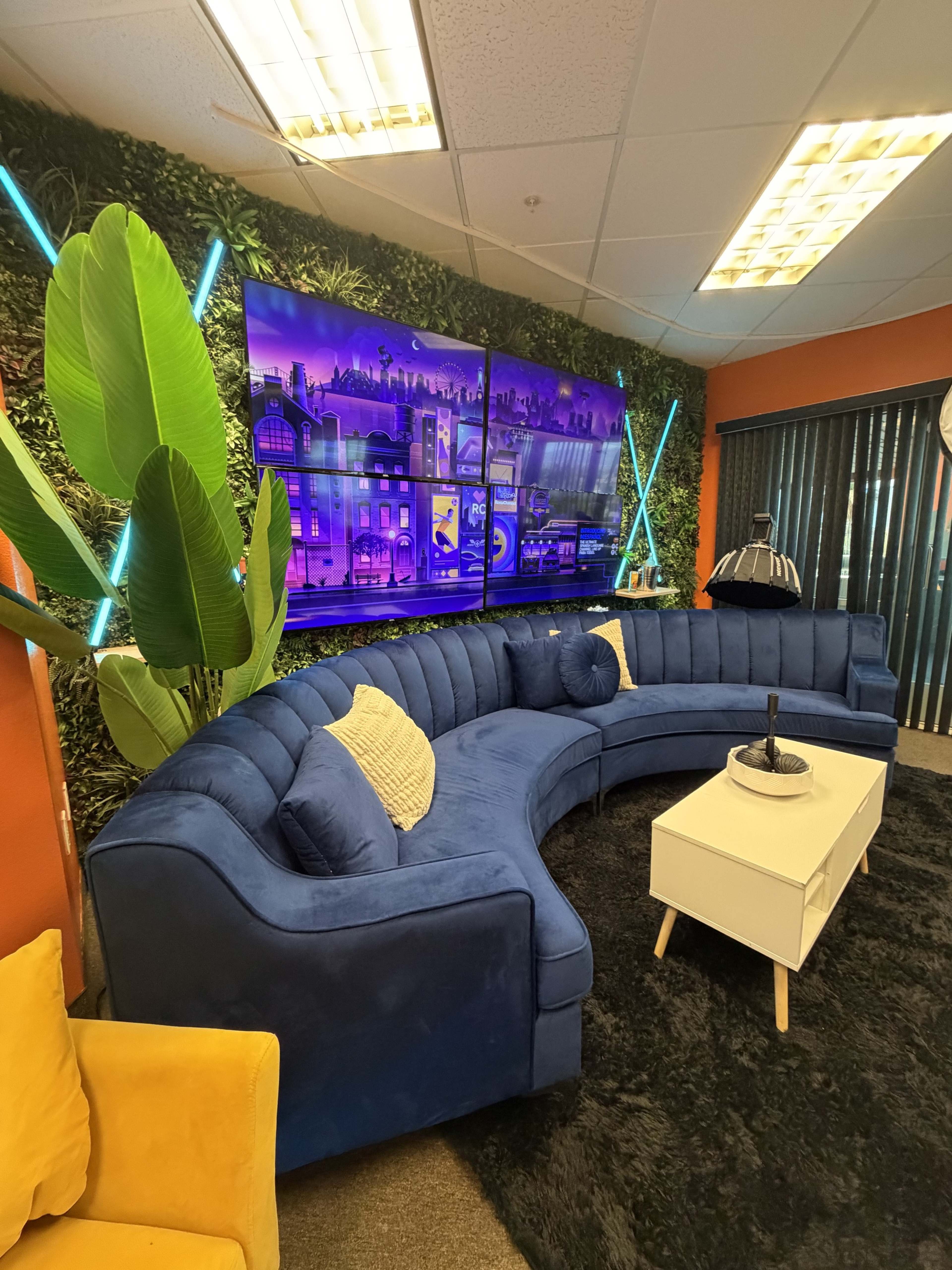 The image shows a modern office lounge area featuring a curved blue couch, a white coffee table, and vibrant wall decor with plants and neon lighting.