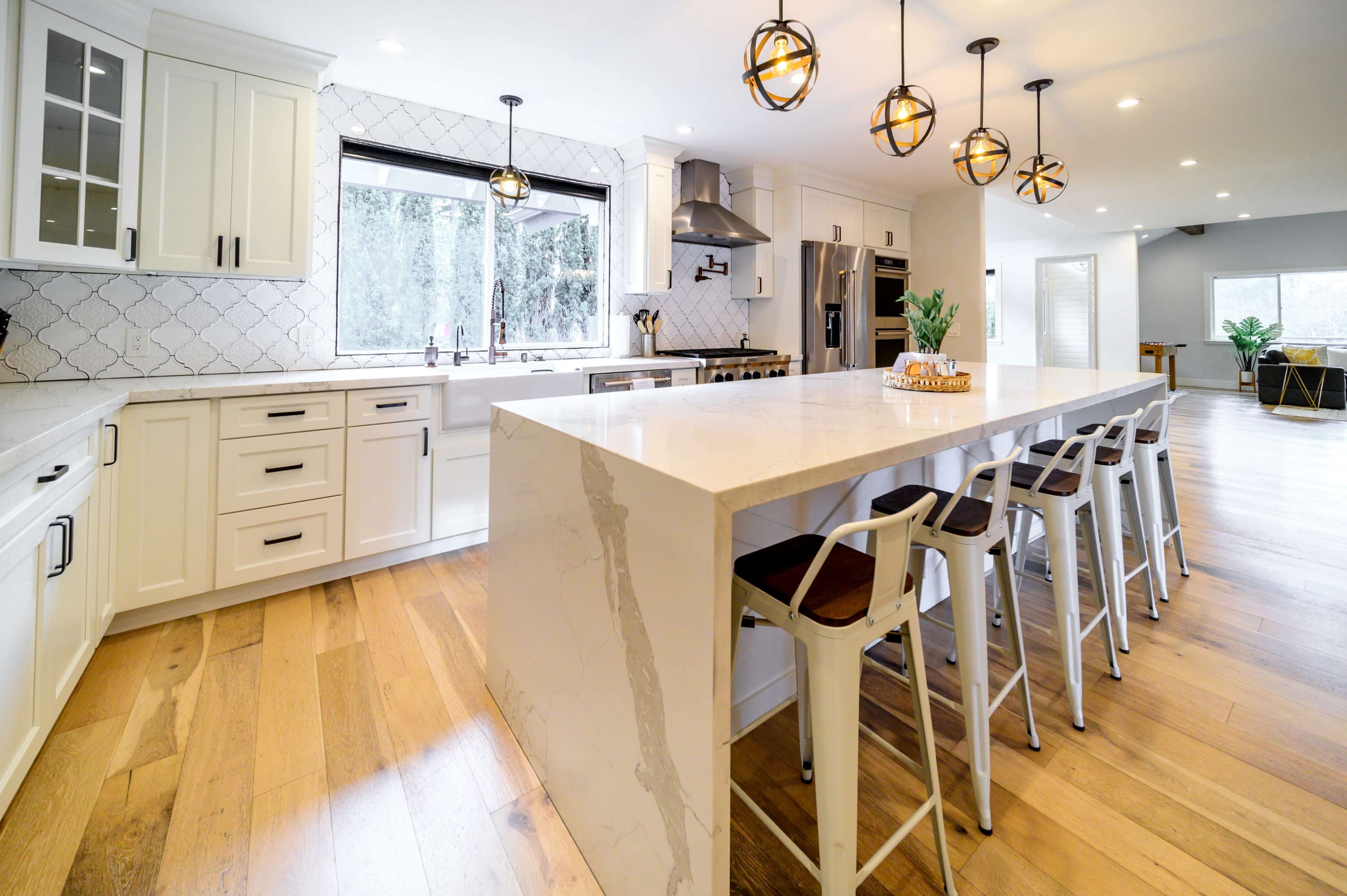 The image shows a modern kitchen with a large island, white cabinetry, and pendant lights, featuring barstools alongside the island.