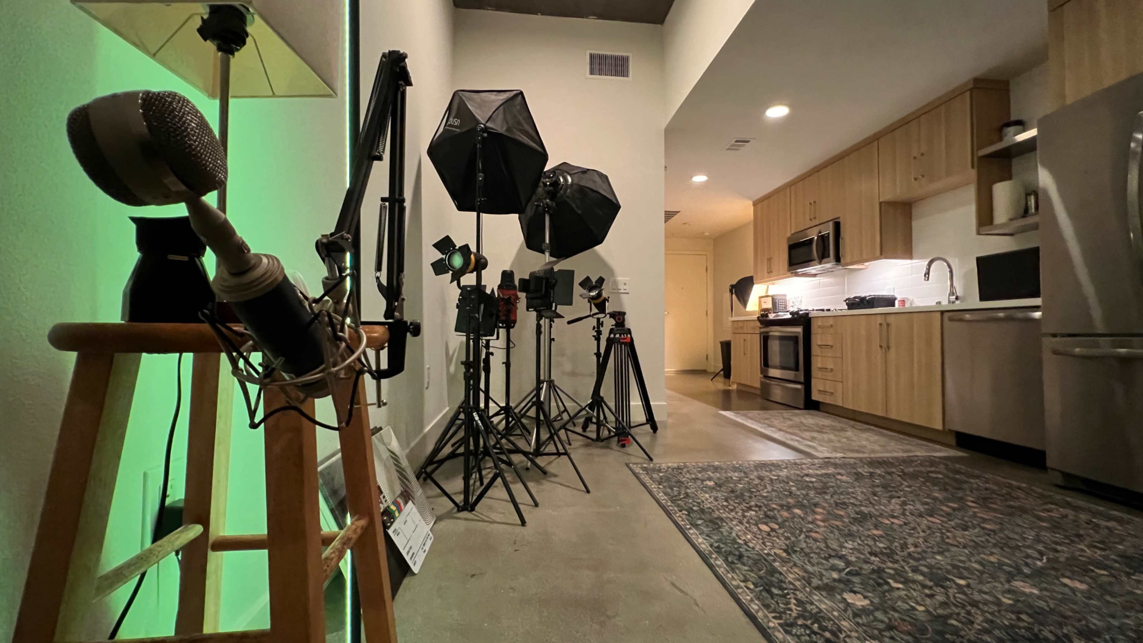 The image shows a modern kitchen setup with various lighting equipment and a microphone stand positioned on a wooden stool.