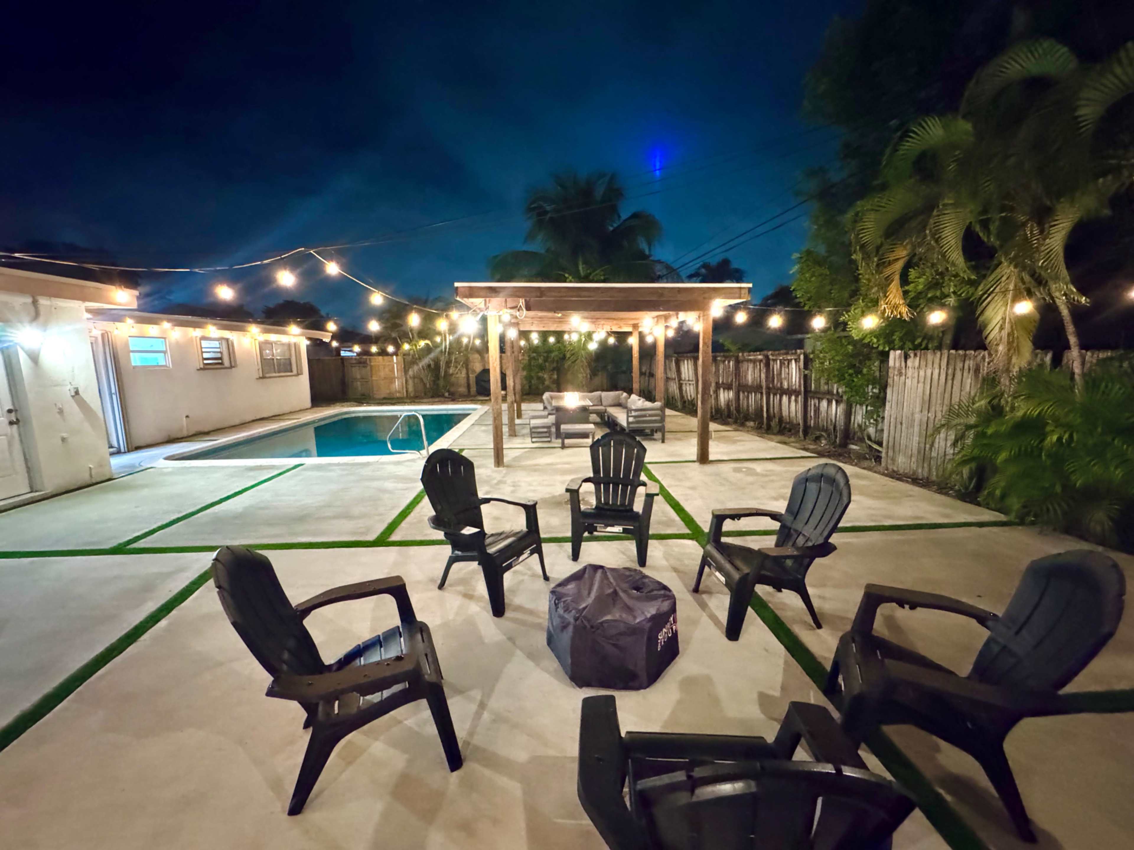 The image shows a nighttime backyard scene with a swimming pool, surrounded by chairs arranged in a circle around a fire pit, and string lights illuminating the area.