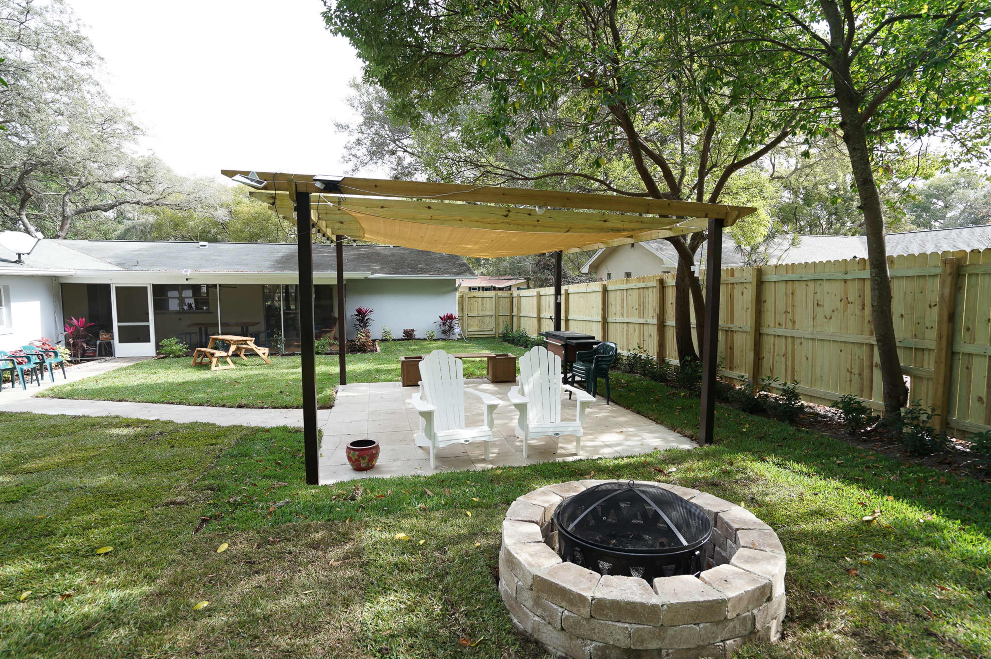 The scene features a backyard patio with a pergola, four white chairs, a grill, and a fire pit surrounded by a stone border, all set against a green lawn and wooden fence.