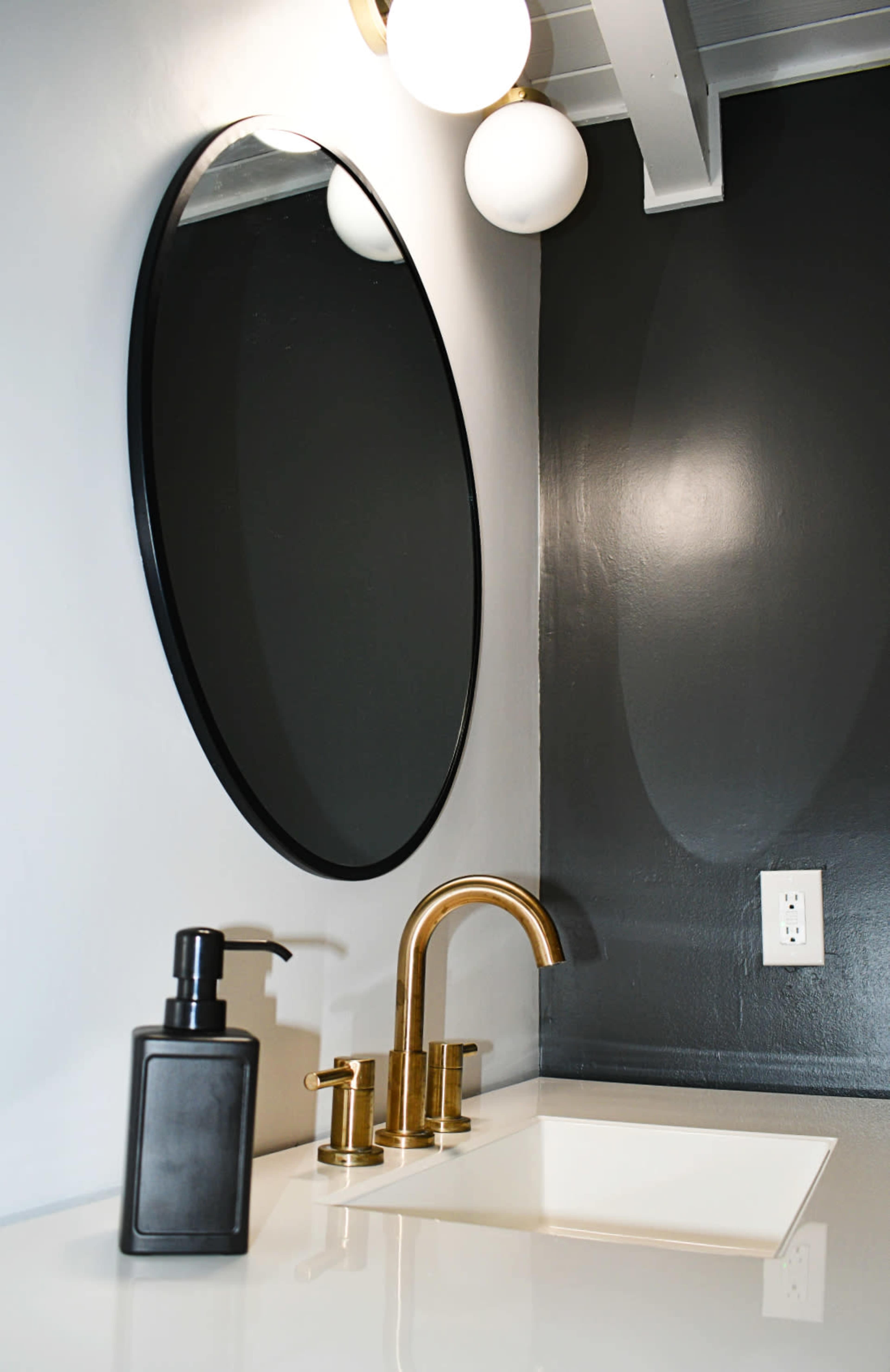 A modern bathroom sink features a circular mirror, a brass faucet, and a black soap dispenser against a dark wall.