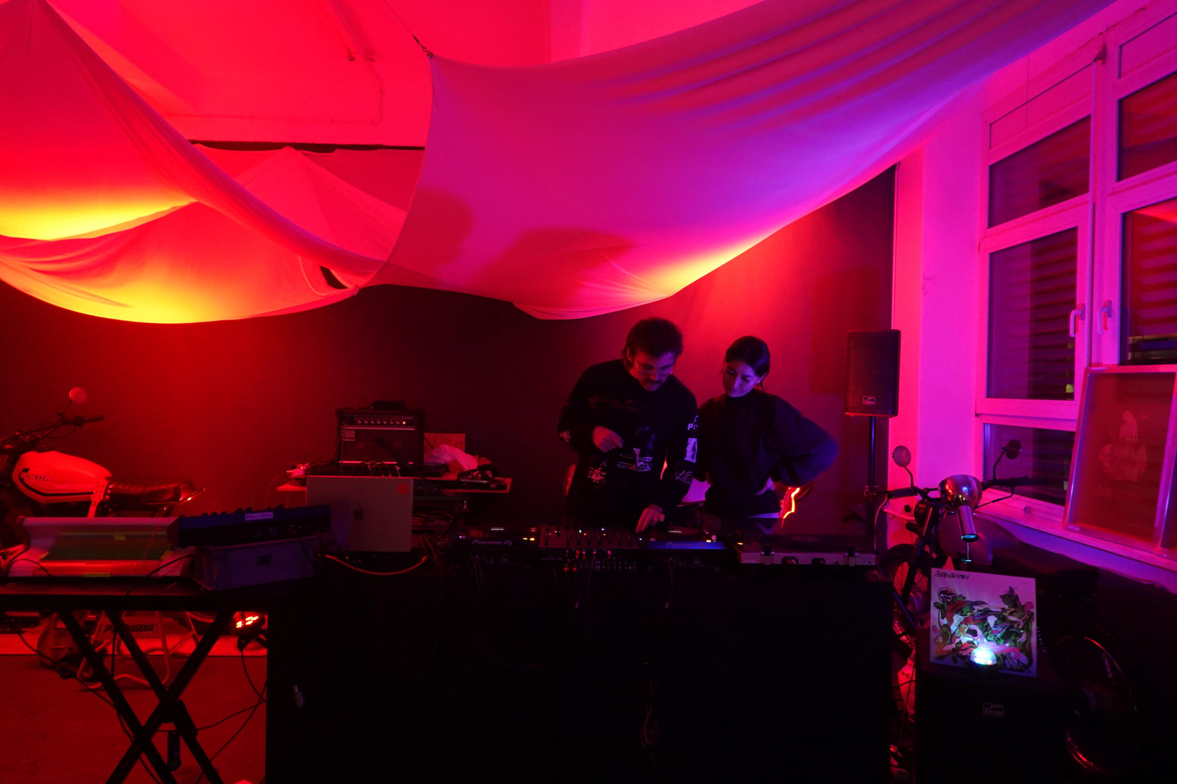Two DJs are performing in a dimly lit room with red lighting and fabric draped from the ceiling, surrounded by electronic equipment and musical instruments.