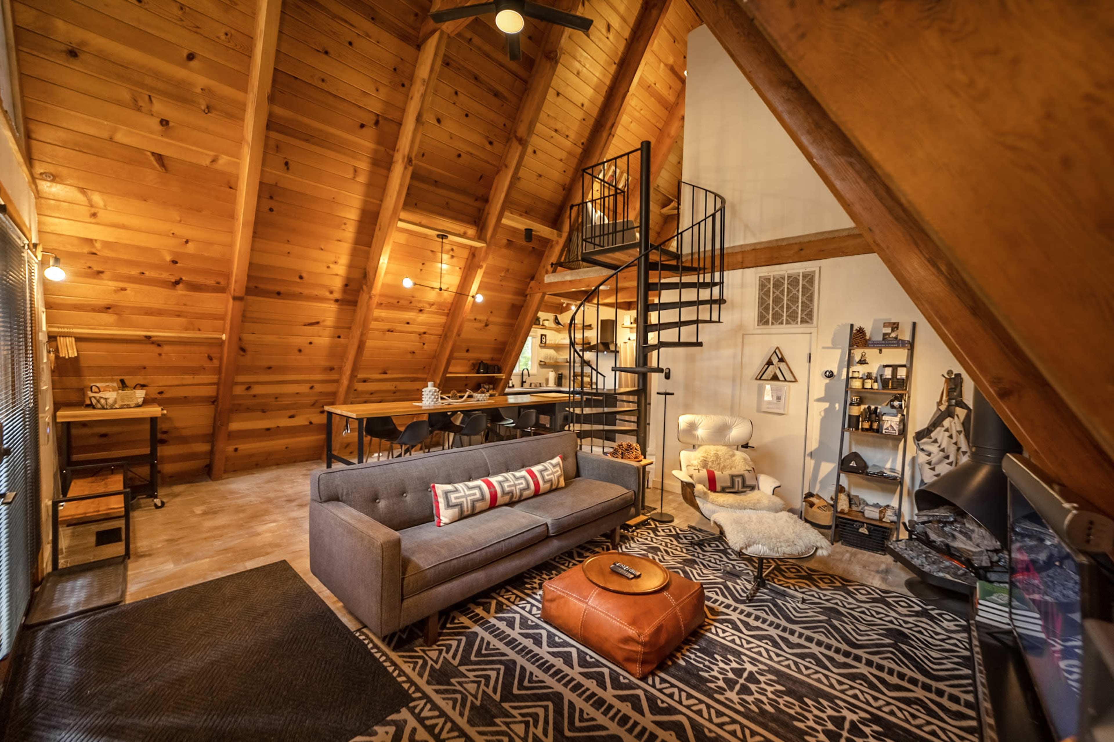 The image depicts a cozy A-frame cabin interior featuring wooden walls, a spiral staircase, a comfortable gray sofa, and a dining area with a large table.