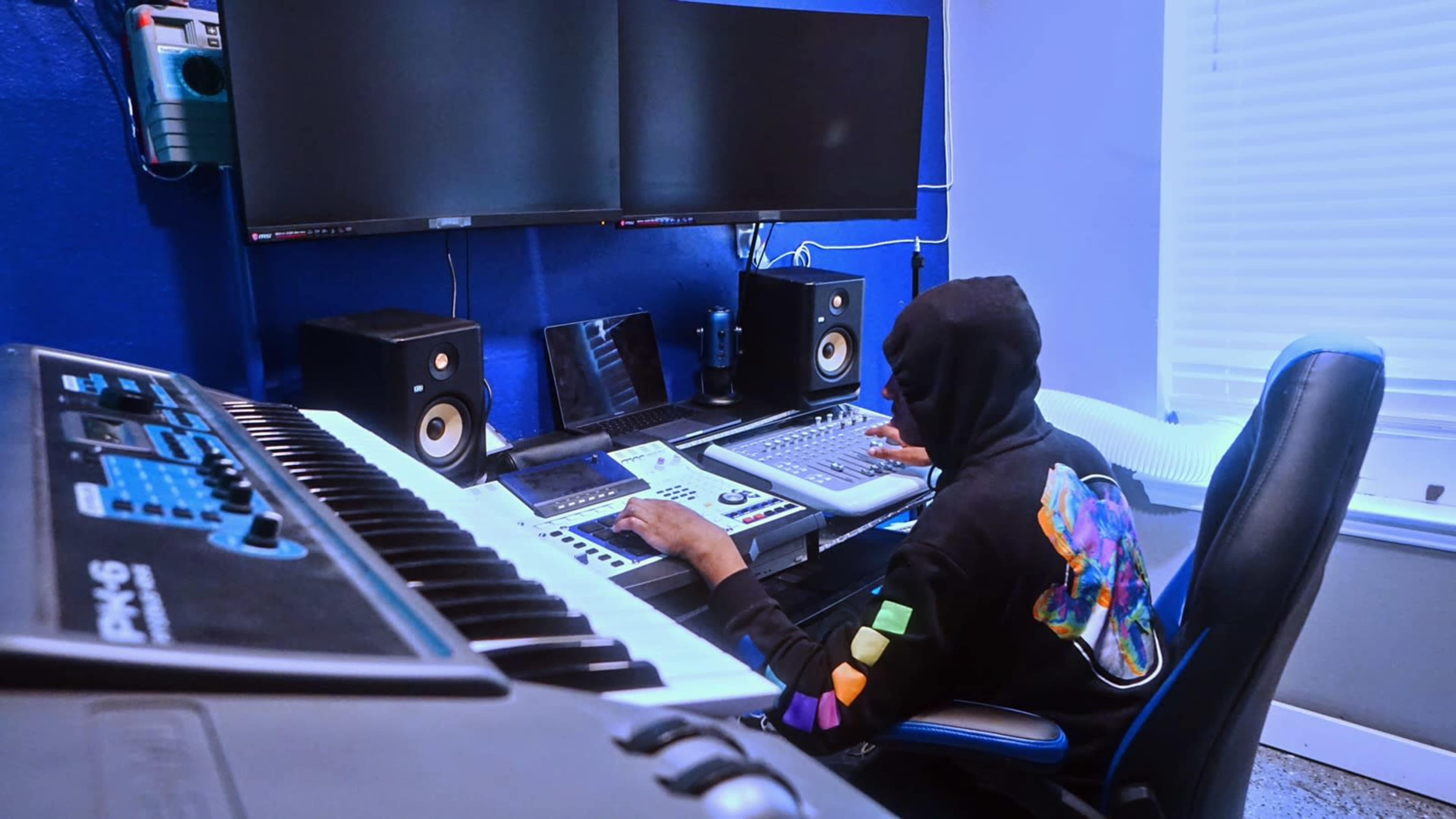 A person wearing a hooded sweatshirt sits at a mixing desk in a music studio, surrounded by computer monitors, speakers, and various audio equipment.