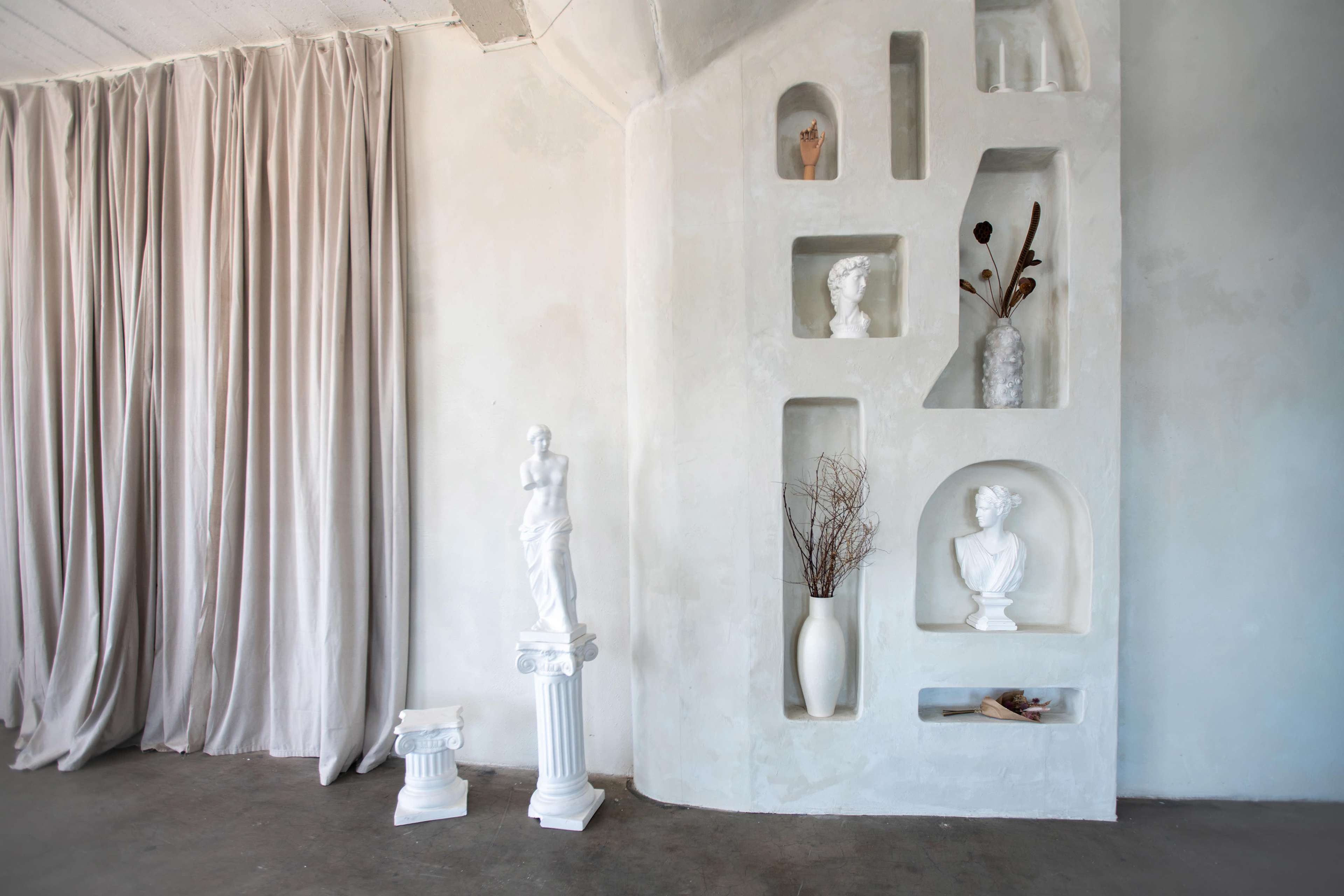 The image shows a minimalist room corner with light-colored walls featuring a niche display of white sculptures and a tall vase, complemented by soft drapes.