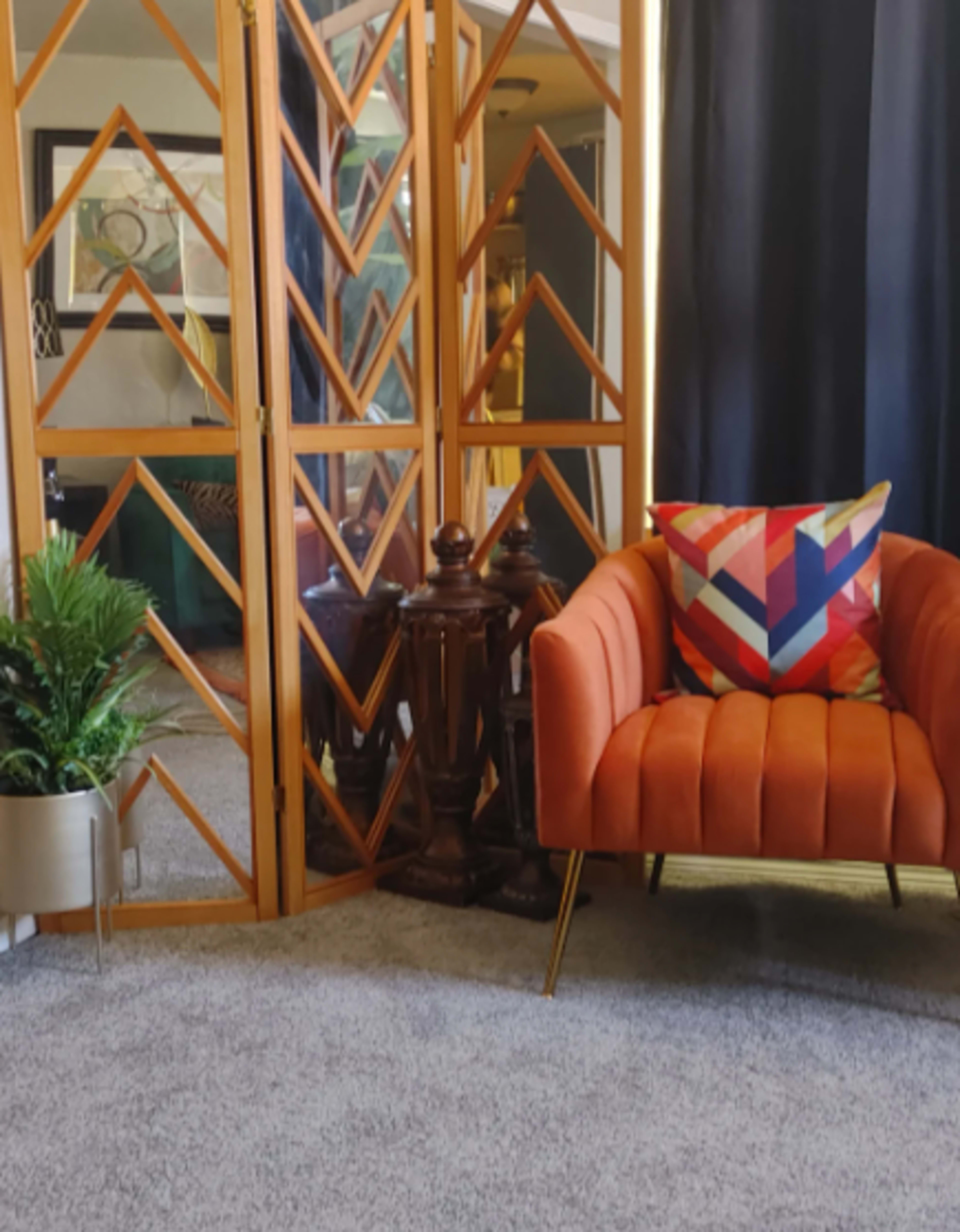 A modern living space features an orange tufted sofa, a geometric pillow, a decorative screen, and a potted plant.