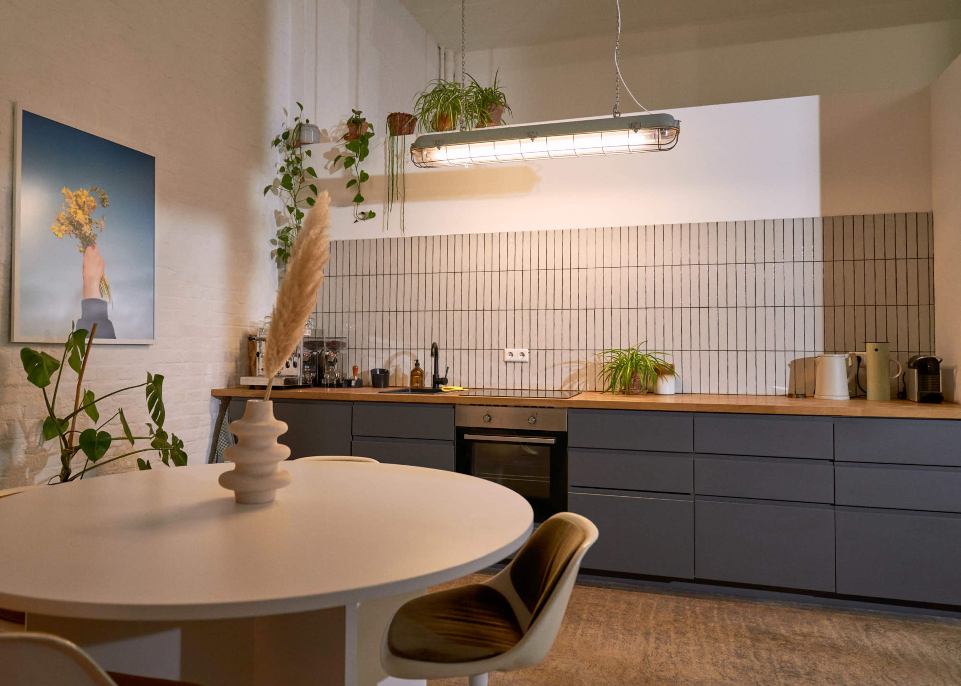 The image shows a modern kitchen with a round dining table, dark cabinets, and a wall of white and gray tiles, illuminated by a linear light fixture.