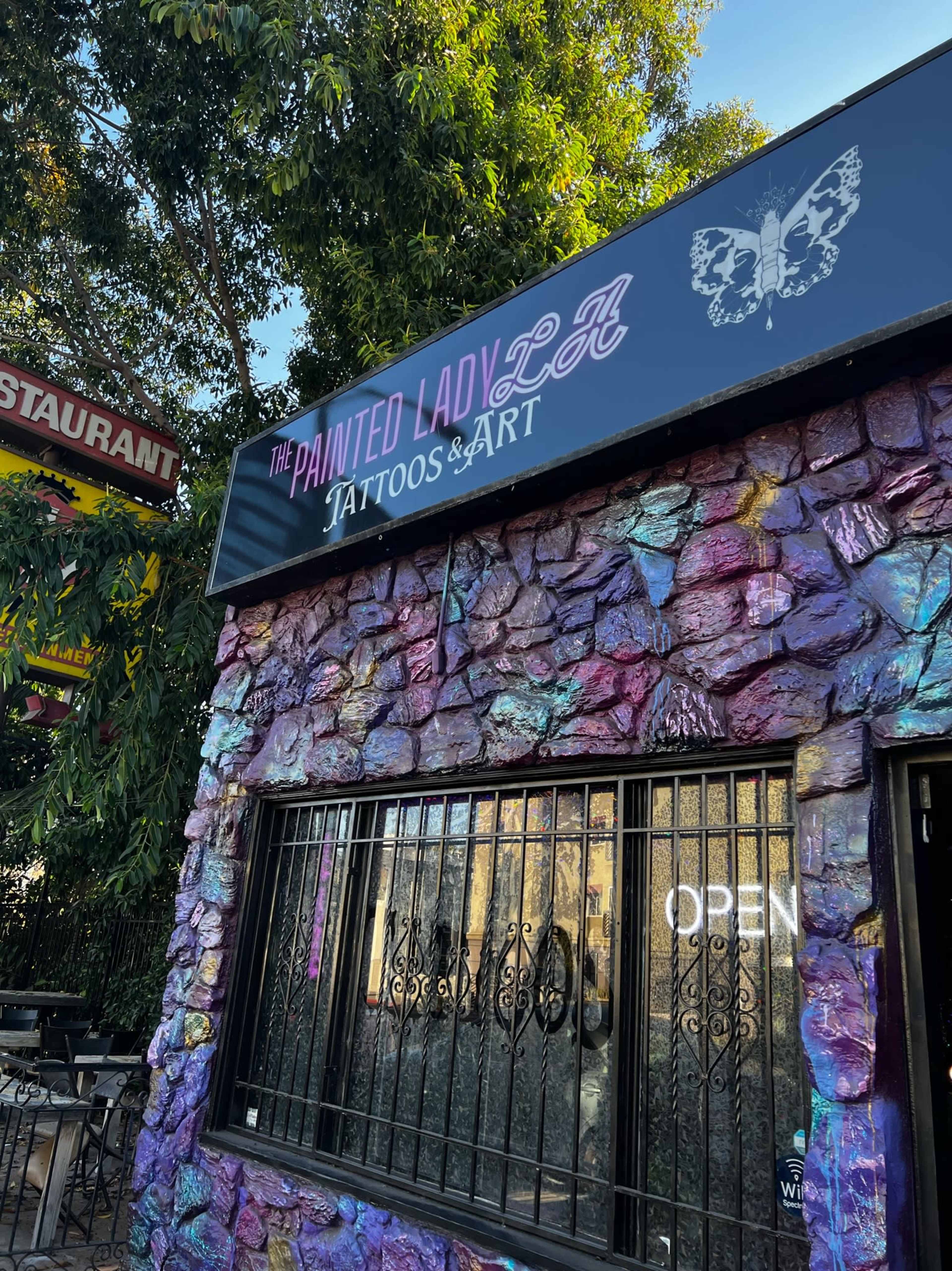 A tattoo shop called "The Painted Lady LA," featuring a colorful stone exterior and a sign with a butterfly motif.