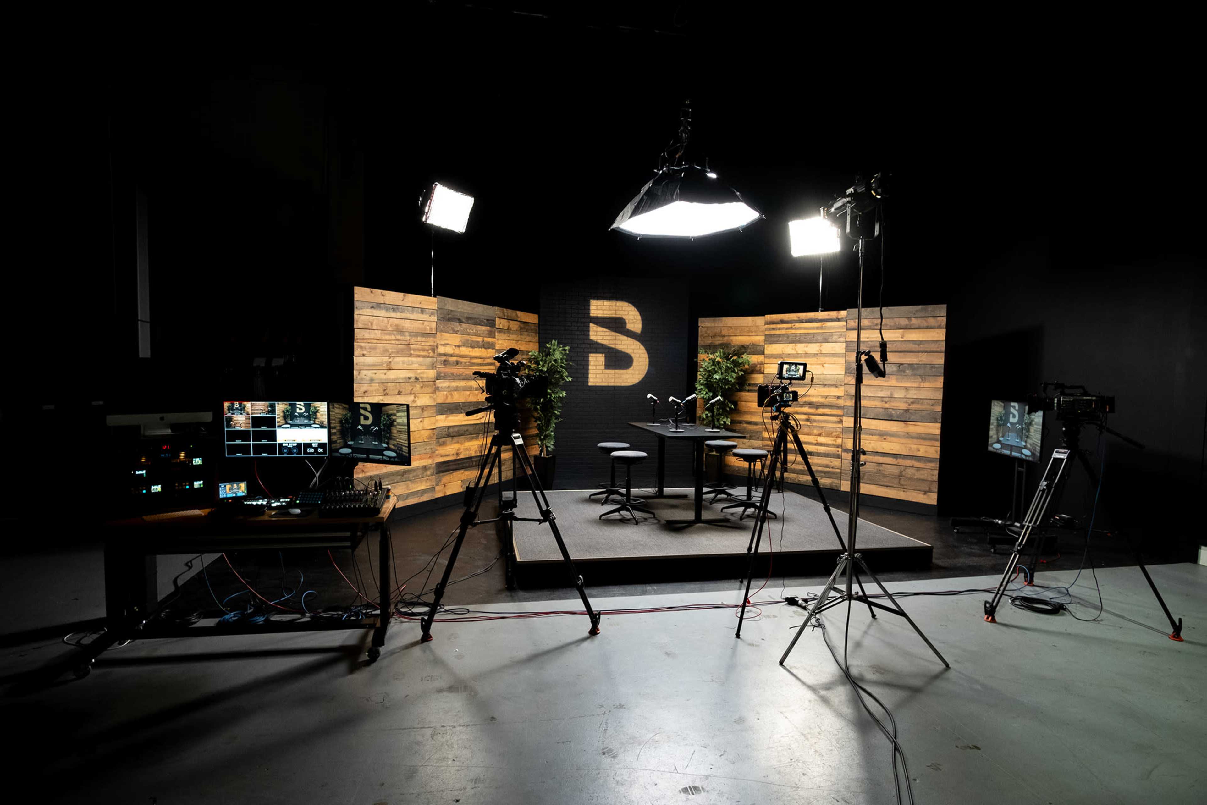 A television studio setup with cameras, lighting equipment, and a wooden backdrop featuring a large letter "S".