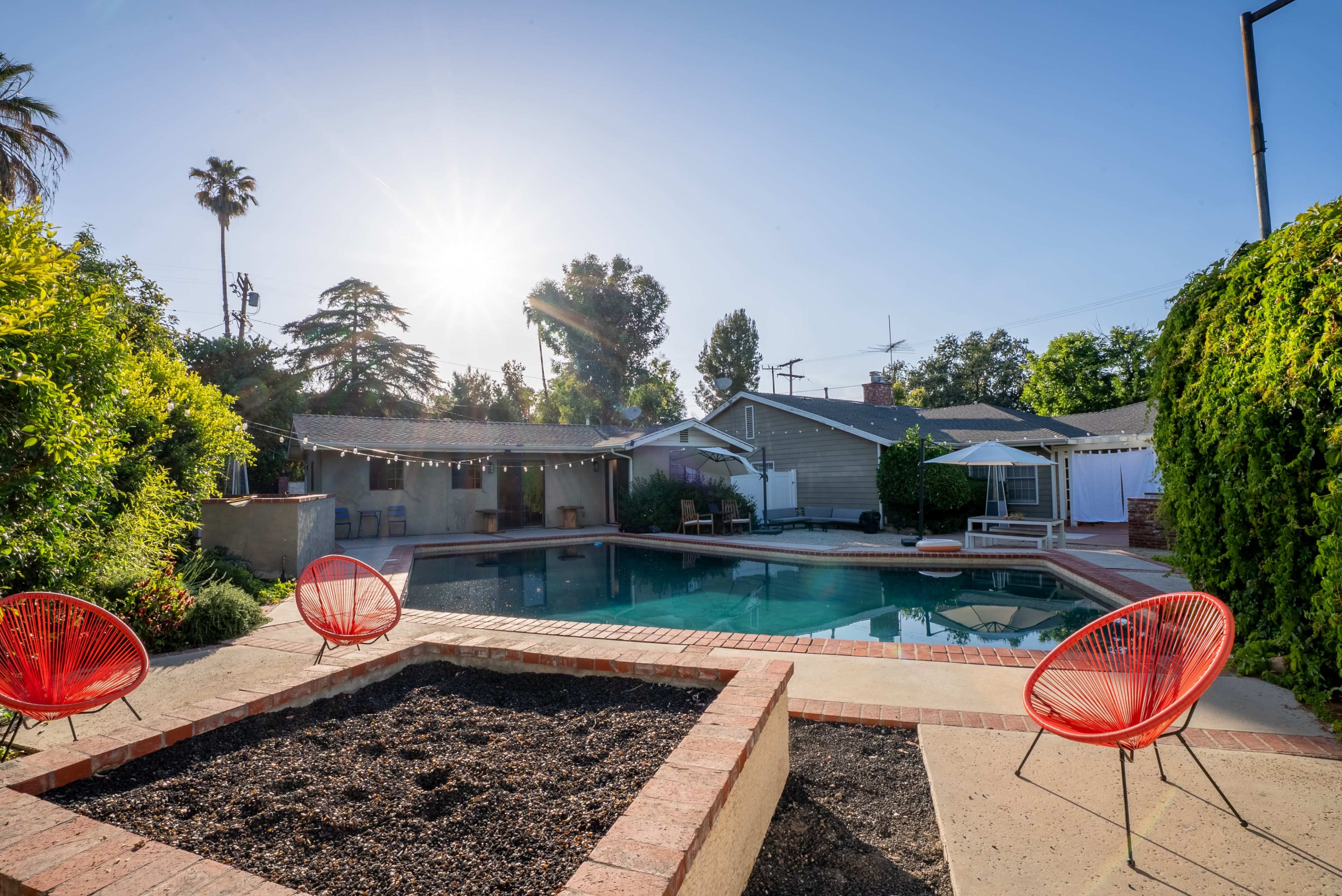 A sunlit backyard features a swimming pool surrounded by a brick patio and three red chairs, with landscaped greenery and houses in the background.