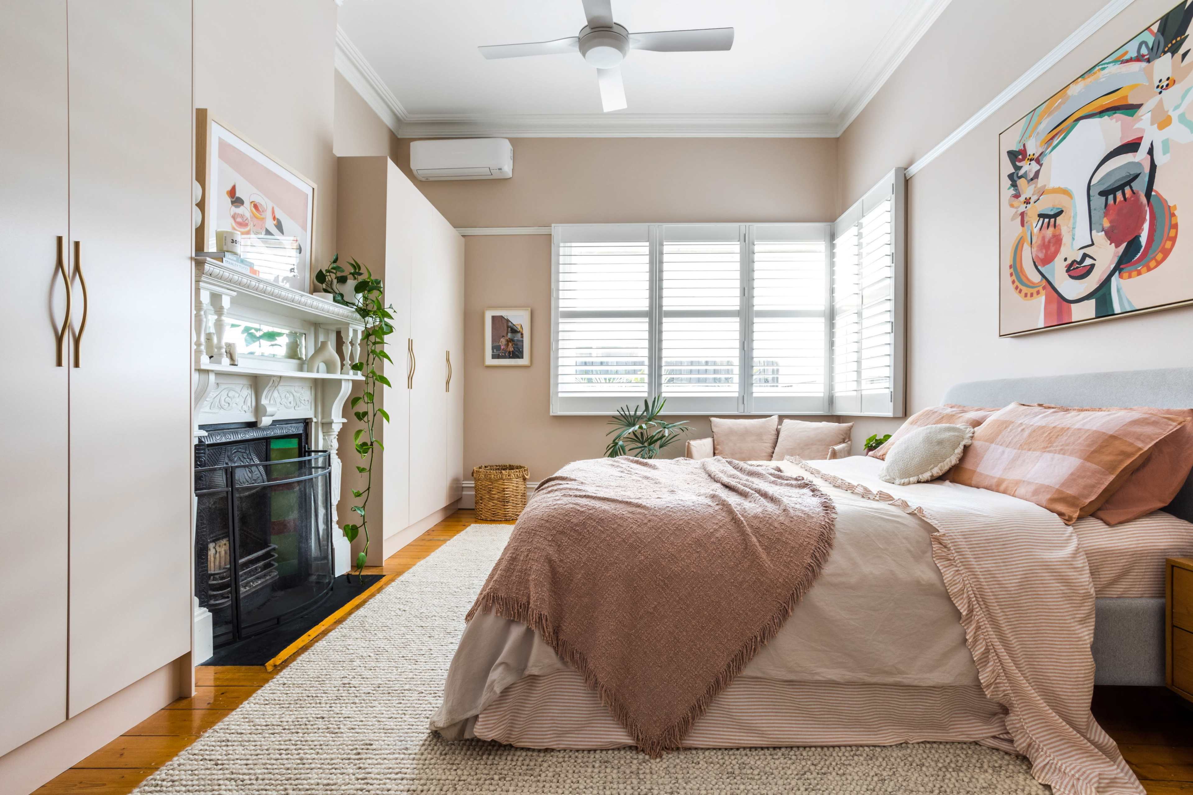 A well-decorated bedroom features a bed with soft linens, a decorative wall art piece, and a fireplace surrounded by plants and a woven basket.