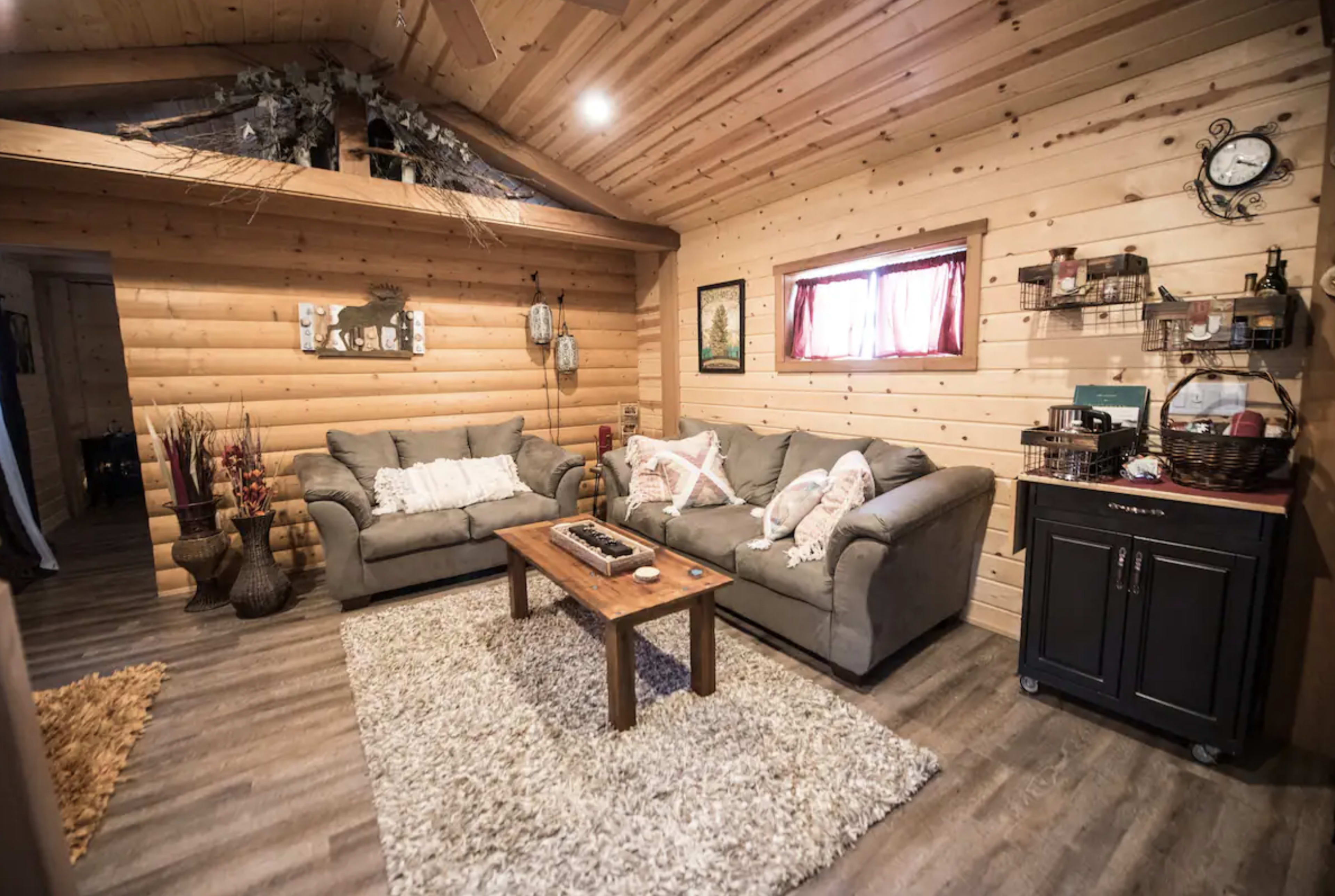 The image shows a cozy living room with wooden walls, two sofas, a coffee table, and a textured rug.