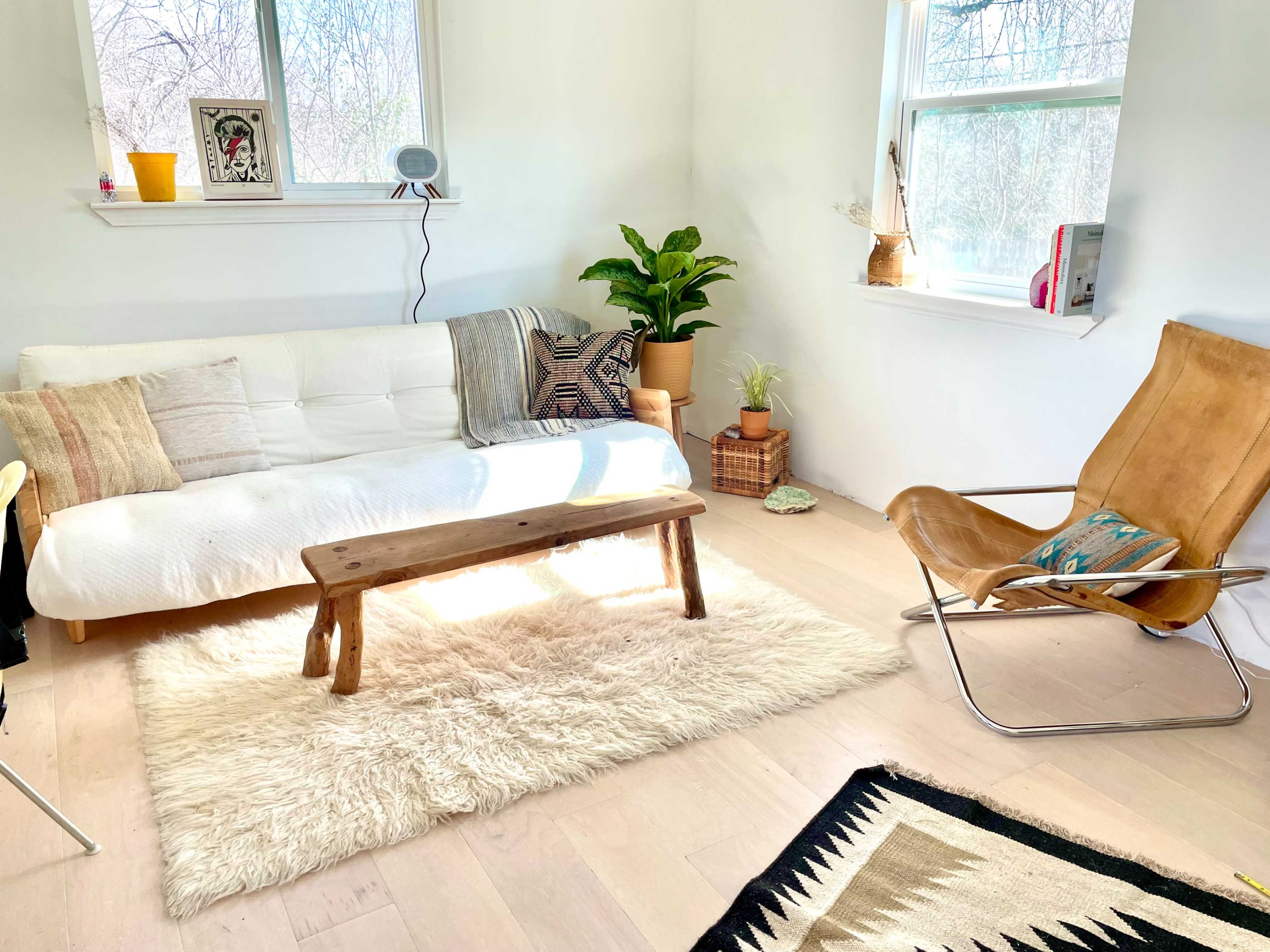 A bright, minimalist living room featuring a white sofa, a wooden coffee table, a beige rug, and a leather chair.