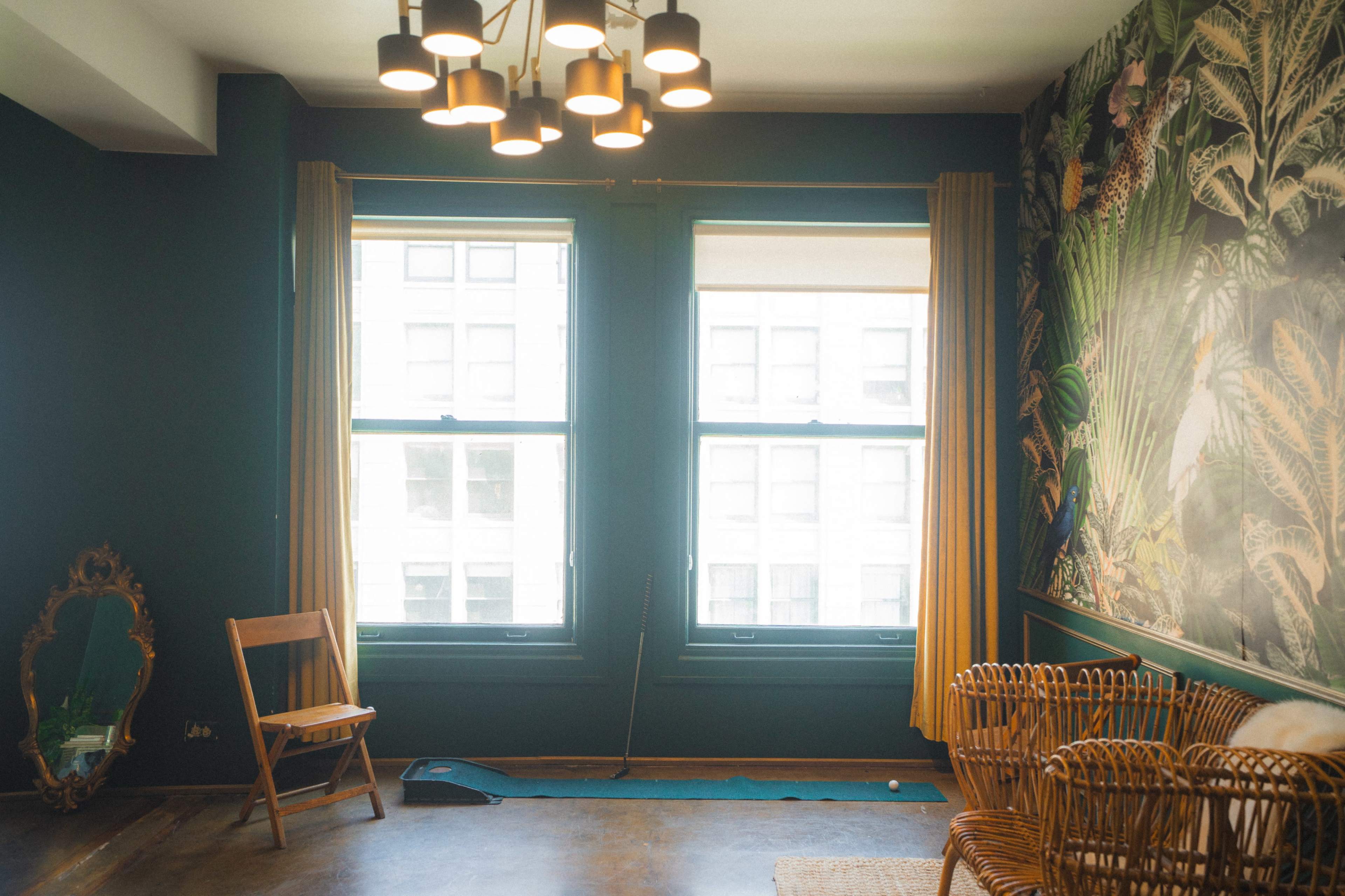 The room features two large windows with green frames, a wicker seating area, a small wooden chair, and a decorative mirror against a wall adorned with tropical wallpaper.