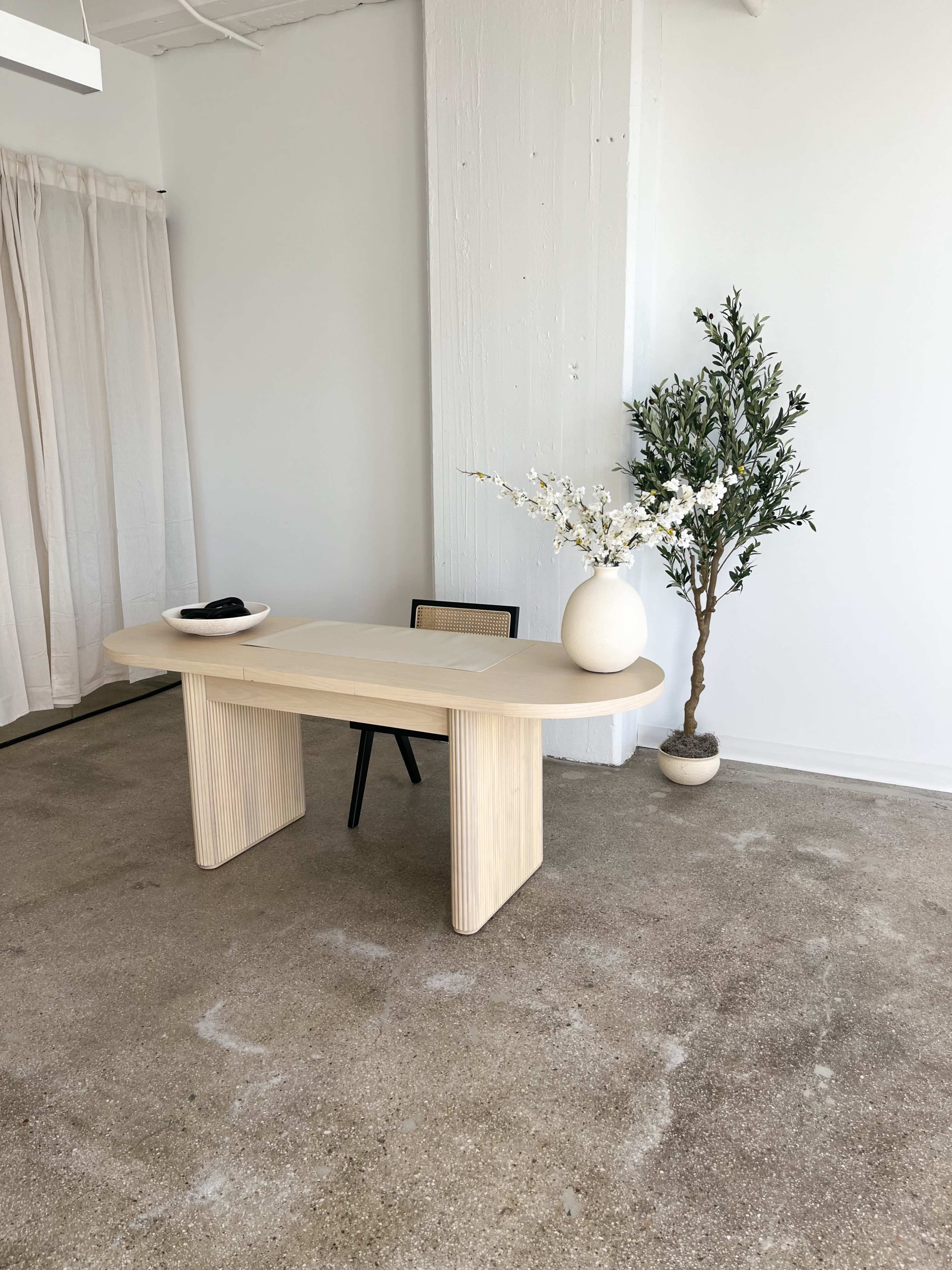 A minimalist office space features a light wooden table with rounded edges, a chair, a vase with branches, and a small plant in a pot.