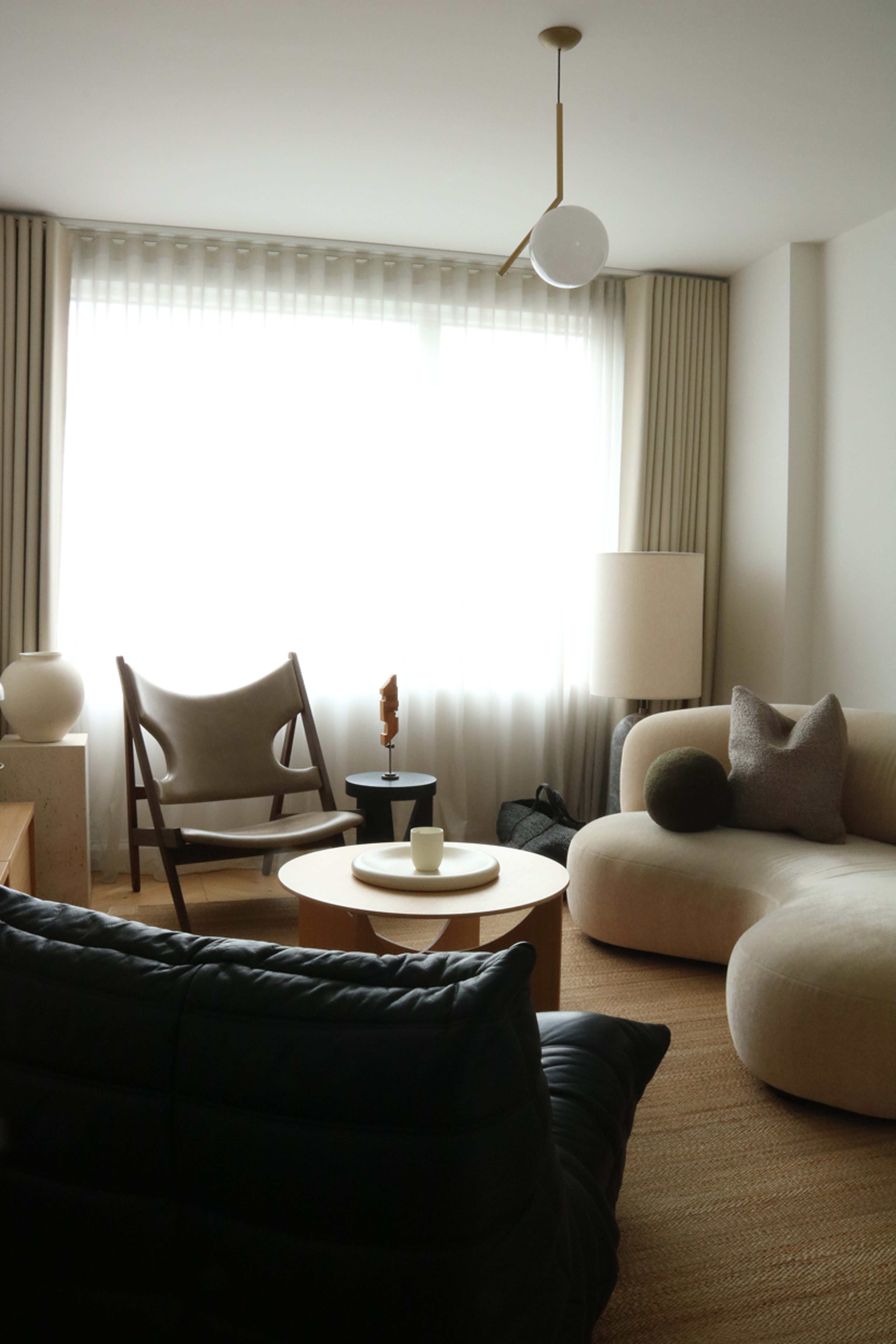 The image shows a modern living room with a circular coffee table, a stylish sofa, a wooden chair, and a large window with sheer curtains letting in soft light.