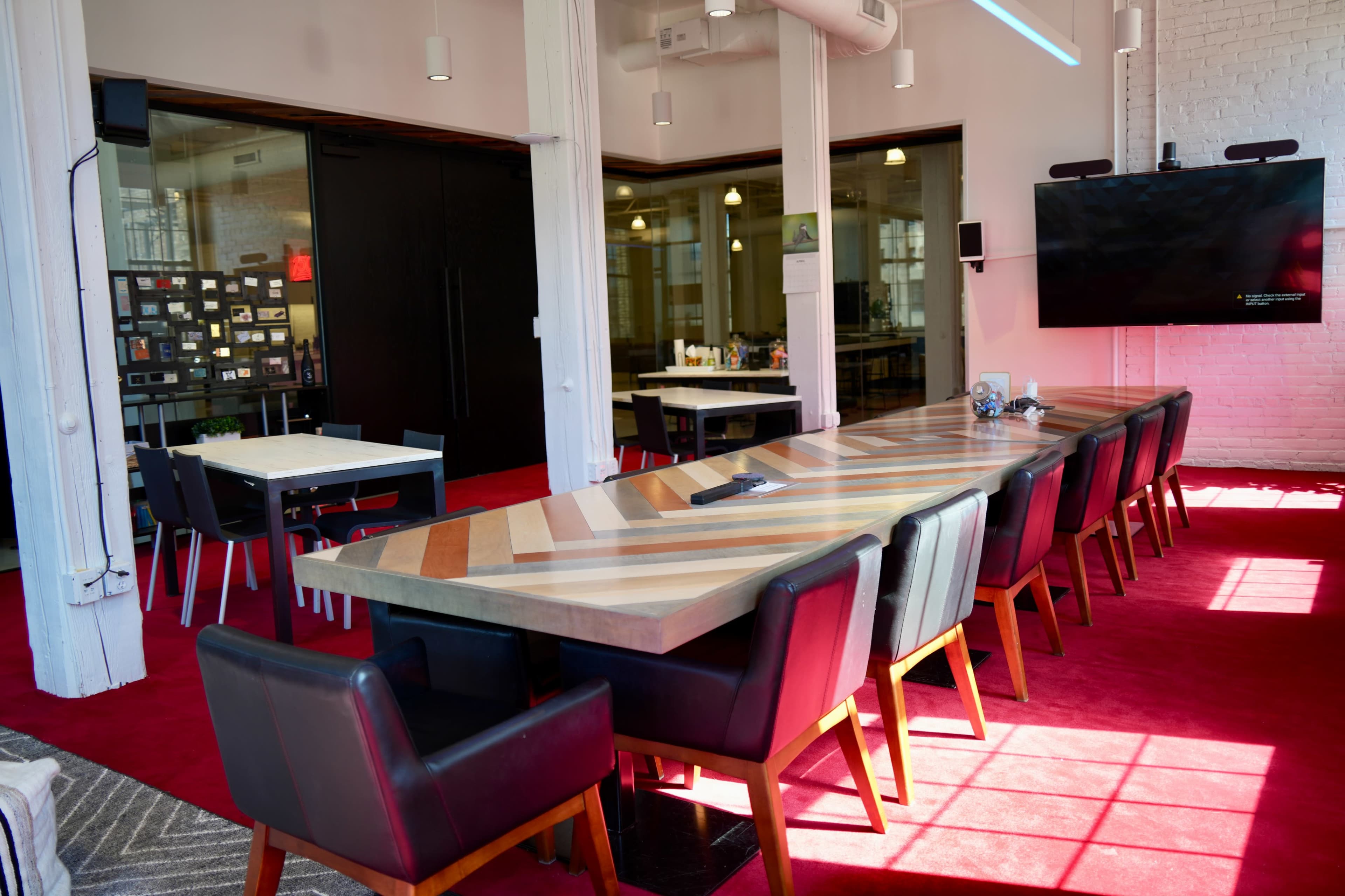 The image shows a modern conference room with a long, striped table, black leather chairs, a large screen mounted on the wall, and a red carpet under bright lighting.