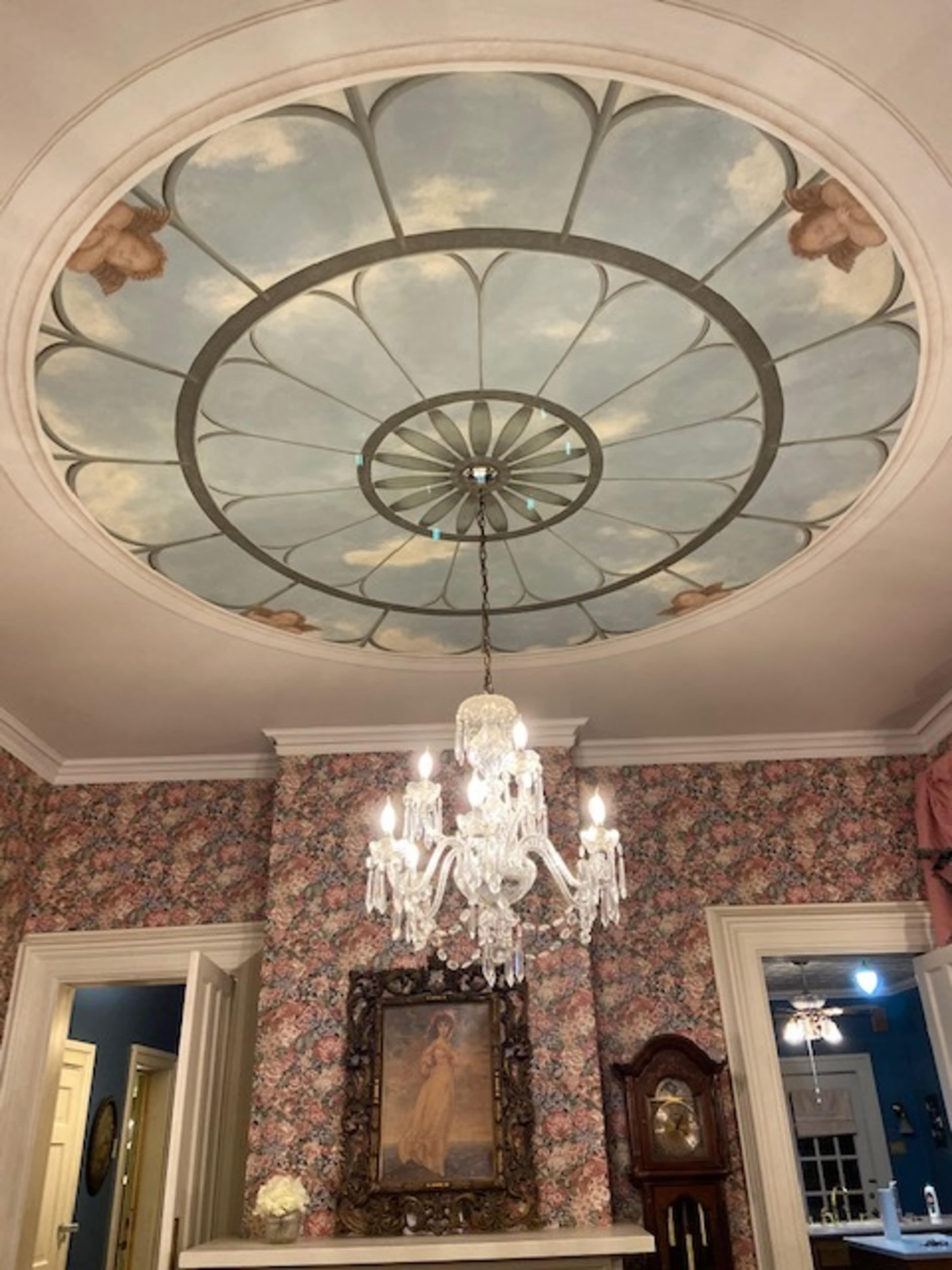 A patterned ceiling with a floral design, adorned with a crystal chandelier, in a room featuring floral wallpaper and an ornate framed painting.